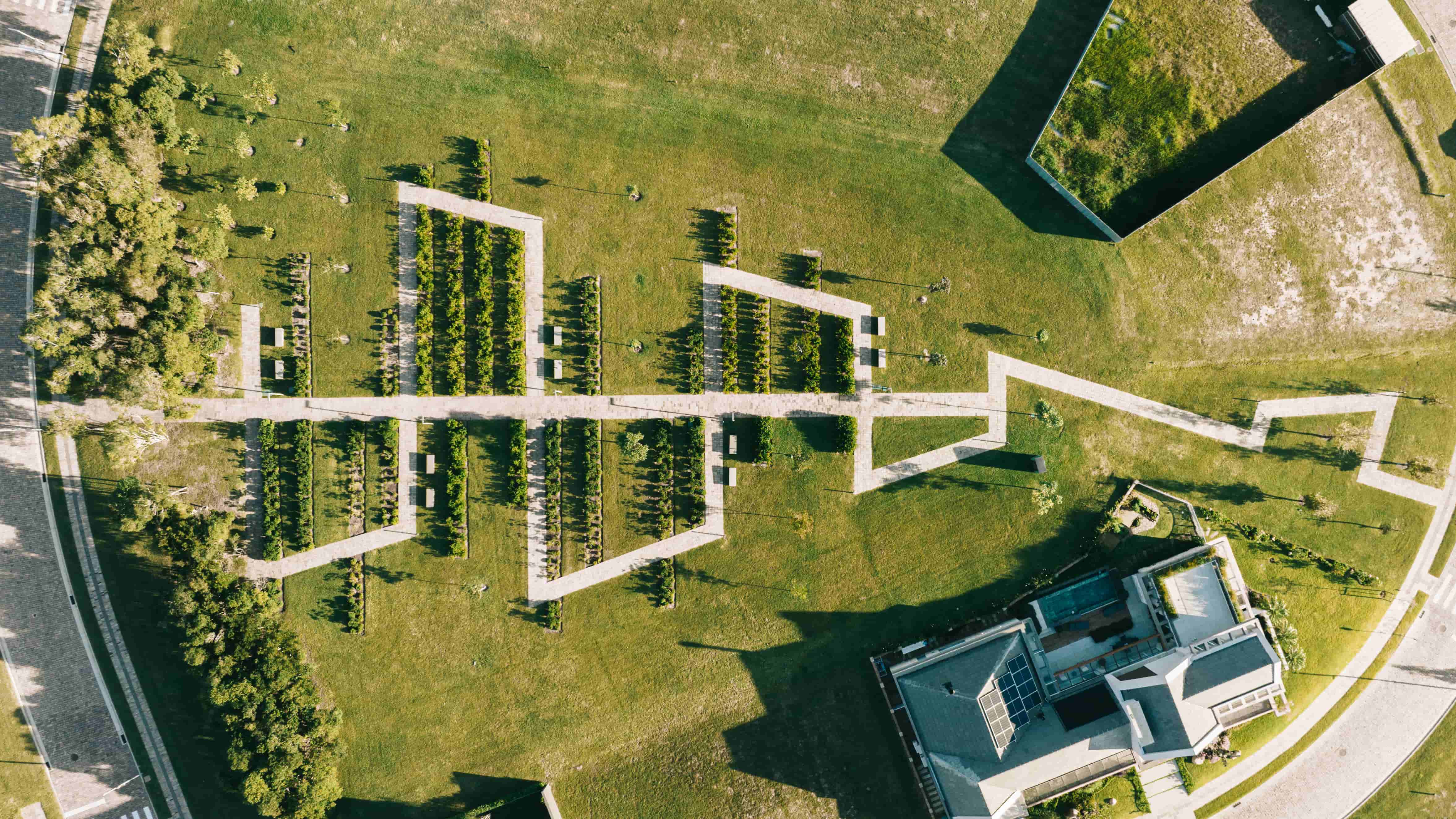 Projeto paisagístico de grande escala com forte linguagem contemporânea, organizado em canteiros geométricos retangulares delimitados por muros baixos de concreto aparente, interligados por uma passarela central elevada que cria um eixo de simetria axial impressionante visto de cima. As espécies arbóreas e arbustivas foram plantadas em fileiras rigorosamente alinhadas dentro dos módulos, criando um ritmo visual que dialoga com a arquitetura das edificações adjacentes — uma delas com cobertura ve, estilo Moderno, elementos Muros baixos de concreto aparente formando canteiros geométricos retangulares, Passarela central elevada em concreto conectando módulos ajardinados, Cobertura verde extensiva em edificação no canto superior direito, Edificação contemporânea com telhado plano e painéis de vidro no canto inferior direito, Plantio em fileiras lineares rigorosamente alinhadas dentro dos canteiros, Via curva asfaltada circundando o conjunto paisagístico, Gramado amplo como pano de fundo entre os canteiros estruturados, Simetria axial no traçado geral do projeto, plantas Murta, Buxinho, Podocarpus, Cipreste-italiano, Grama-esmeralda, Jabuticabeira, Ipê, Palmeira-real, Cerca-viva de Viburno, Ligustro, ambiente moderno, sofisticado, localização jardim, entrada, corredor, área grande, estruturas iluminacao, banco, cores verde-escuro, verde-claro, cinza-concreto, bege, branco, marrom, piso grama, pedra, porcelanato, privacidade cerca_viva, muro_verde, água nenhum