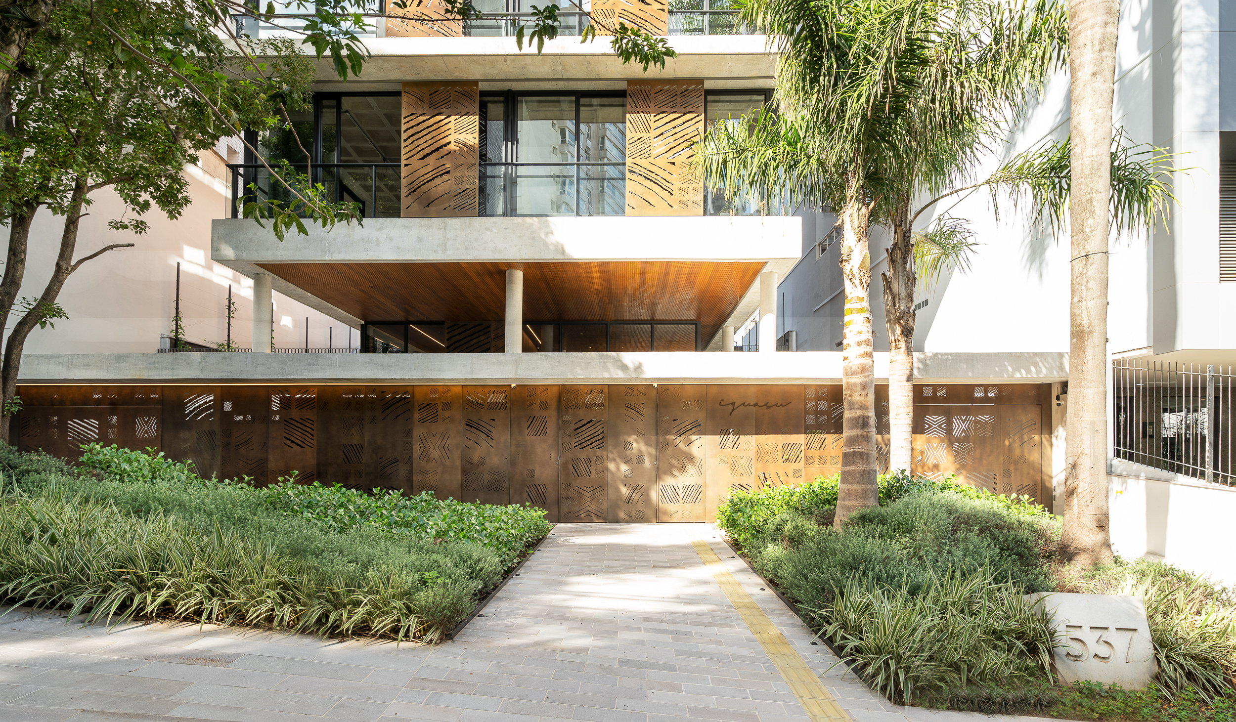 Projeto paisagístico contemporâneo de entrada de edifício residencial de alto padrão em São Paulo, com canteiros simétricos e generosos de forração composta por amendoim-rasteiro e barba-de-serpente que emolduram o passeio central em pedra natural, criando um percurso convidativo até o lobby com marquise em concreto aparente e forro de madeira. A fachada é marcada por painéis de aço corten perfurado com grafismos diagonais que dialogam com a vegetação tropical, incluindo palmeiras-reais de porte, estilo Moderno, Tropical, elementos Painéis metálicos perfurados em aço corten com padrões geométricos diagonais na fachada e muro frontal, Laje de concreto aparente em balanço formando marquise, Forro em madeira ripada sob a marquise, Pilares cilíndricos em concreto aparente, Piso em placas de pedra natural tipo São Tomé ou ardósia em tons cinza, Passeio central em paginação retilínea com pedras de formato retangular, Marco em pedra natural com numeração 537, Esquadrias em alumínio preto com vidro, Gradil metálico lateral em barras verticais, Letreiro metálico cursivo na fachada (Iguaçu), plantas Amendoim-rasteiro, Barba-de-serpente, Liriope, Palmeira-real, Jabuticabeira ou árvore de médio porte não identificada, Salsa-chorão ou Chorão-japonês, Buxinho, Moréia, Pingo-de-ouro, ambiente moderno, sofisticado, localização entrada, fachada, jardim, área medio, estruturas iluminacao, cores Verde escuro, Verde claro, Corten/ferrugem, Cinza concreto, Madeira natural, Preto, Bege, piso pedra, pisantes, privacidade cerca_viva, água nenhum