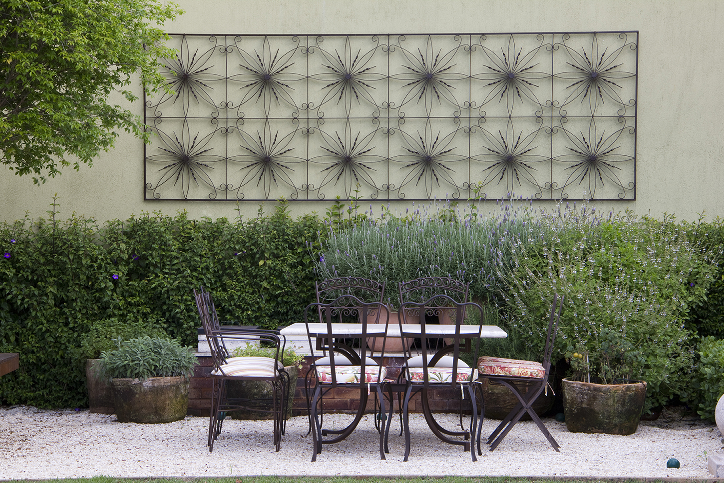 Jardim de inspiração provençal com atmosfera europeia, onde um elegante painel de ferro forjado com motivos florais repetitivos adorna o muro bege, servindo como ponto focal sobre uma massa exuberante de lavandas em plena floração lilás e cerca viva de murta rigorosamente podada. O espaço de convívio é composto por um conjunto de mesa e cadeiras em ferro forjado com almofadas estampadas, posicionado sobre piso de brita branca, ladeado por vasos de barro rústicos e canteiros elevados em tijolos c, estilo Classico, elementos Painel decorativo em ferro forjado com motivos florais fixado no muro, Mesa de jantar em ferro forjado com tampo escuro, Conjunto de 4 cadeiras em ferro forjado com almofadas estampadas florais e lisas, Vasos de barro envelhecidos de diferentes tamanhos, Canteiro elevado em tijolos aparentes, Muro rebocado em tom bege/verde claro, Cerca viva podada em formato retangular, plantas Lavanda, Murta, Alecrim, Hortelã ou erva aromática não identificada, Árvore de médio porte não identificada, ambiente relaxante, sofisticado, acolhedor, localização quintal, muro, área medio, estruturas banco, iluminacao, cores Verde escuro, Verde acinzentado, Lilás/lavanda, Preto fosco (ferro), Bege claro, Branco (almofadas), Terracota (vasos), Marrom (tijolos), piso brita, privacidade cerca_viva, muro_verde, água nenhum