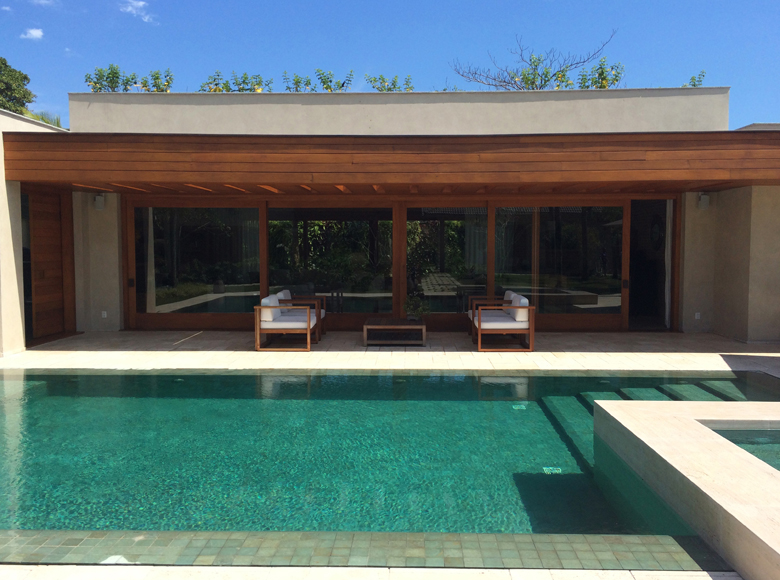 Projeto paisagístico contemporâneo de alto padrão que integra harmoniosamente a arquitetura minimalista com vegetação tropical exuberante, destacando-se a piscina em tom esmeralda com spa integrado e bordas em pedra calcária natural. O pergolado em madeira cumaru com painéis de vidro piso-teto cria uma transição fluida entre o interior e o exterior, enquanto o teto verde na cobertura e a massa vegetal ao fundo proporcionam conforto térmico e privacidade. A composição paisagística utiliza espécie, estilo Moderno, Tropical, elementos Piscina retangular em tom esmeralda com borda infinita, Spa/jacuzzi integrada à piscina com degraus submersos, Deck de pedra natural calcária ao redor da piscina, Pergolado de madeira cumaru com cobertura parcial, Pórtico com colunas de madeira e vidro piso-teto, Mobiliário de design com poltronas em madeira e estofado branco, Mesa de centro em madeira maciça, Teto verde/jardim na laje da cobertura, Painéis de vidro deslizantes de grandes dimensões, Muros em concreto aparente com acabamento cimentício, plantas Clúsia, Palmeira-ráfis, Bambu-mossô, Helicônia, Jabuticabeira, Ficus-benjamina, Grama-esmeralda, ambiente sofisticado, relaxante, moderno, localização piscina, jardim, varanda, área grande, estruturas pergolado, iluminacao, banco, cores verde-esmeralda, madeira-cumaru, bege-claro, branco, cinza-concreto, azul-céu, piso pedra, deck, grama, privacidade cerca_viva, muro_verde, água piscina