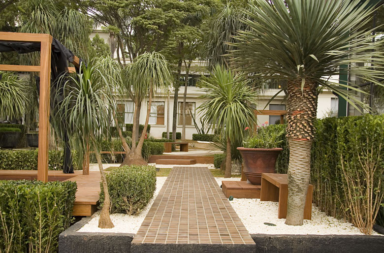 Jardim de estilo contemporâneo-tropical com forte influência do paisagismo brasileiro, estruturado a partir de um caminho central em piso de tijolinho cerâmico ladeado por pedrisco branco que confere leveza e luminosidade ao conjunto. Patas-de-elefante com troncos escultóricos e dracenas conferem verticalidade e exotismo, enquanto topiarias de buxinho em forma de bola garantem a geometria e o rigor formal do projeto. A composição é arrematada por um pergolado de madeira com deck elevado, bancos , estilo Moderno, Tropical, elementos Piso em tijolinho cerâmico retangular formando caminho central, Pedrisco branco como forração de piso, Bancos de madeira maciça em design contemporâneo, Estrutura tipo pergolado em madeira de lei com cobertura preta retrátil, Deck de madeira elevado junto ao pergolado, Vaso grande de barro/cerâmica com palmeira-azul, Mureta de contenção em pedra escura na entrada, Topiaria geométrica em buxinho, plantas Pata-de-elefante, Palmeira-azul, Dracena, Buxinho, Kaizuka, Tuia-holandesa, Pinheiro-negro-japonês, ambiente sofisticado, moderno, relaxante, localização jardim, entrada, corredor, área medio, estruturas pergolado, banco, cores verde-escuro, verde-claro, branco, marrom, terracota, cinza, piso pisantes, brita, deck, privacidade cerca_viva, água nenhum