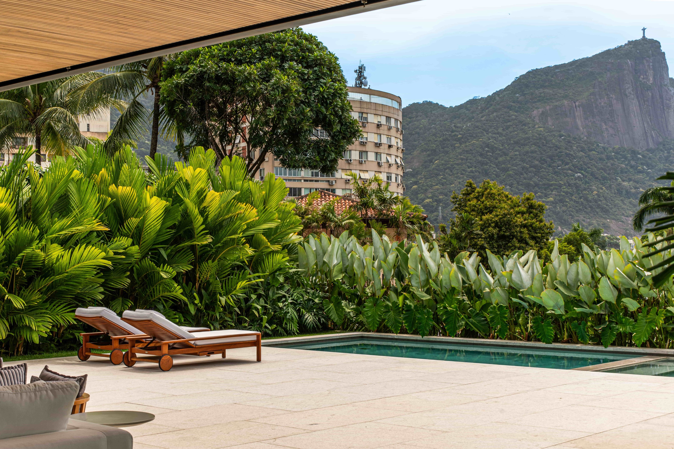 Projeto paisagístico tropical contemporâneo em residência de alto padrão na zona sul do Rio de Janeiro, com vista privilegiada para o Cristo Redentor no Morro do Corcovado. O jardim é composto por maciços densos de vegetação tropical — incluindo palmeiras-areca, estrelítzias, helicônias e filodendros — que funcionam como barreira verde de privacidade ao redor da piscina retangular de borda infinita com acabamento em pedra clara. A área de lazer é marcada por linhas minimalistas no mobiliário de , estilo Moderno, Tropical, elementos Piscina retangular com borda em pedra clara, Duas espreguiçadeiras de madeira teca com estofado branco e rodízios, Sofá modular cinza com almofadas decorativas, Mesa lateral redonda cinza, Cobertura plana com forro de madeira ripada (pergolado contemporâneo), Piso de pedra natural clara em grandes placas, Maciço vegetal tropical como cerca-viva perimetral, Vista para o Cristo Redentor e Morro do Corcovado, plantas Palmeira-areca, Helicônia, Bananeira-ornamental, Coqueiro, Filodendro, Estrelítzia-branca / Bananeira-do-brejo, Calathea, Palmeira-leque, Mangueira, Alpínia, ambiente relaxante, moderno, sofisticado, localização piscina, jardim, varanda, área grande, estruturas pergolado, cores Verde-escuro, Verde-claro, Branco, Bege, Cinza-claro, Marrom-madeira, Azul-piscina, piso pedra, grama, privacidade cerca_viva, muro_verde, água piscina