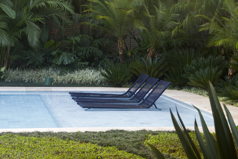 Projeto paisagístico tropical contemporâneo que emoldura uma piscina retangular com bordas suavizadas em pedra São Tomé branca, criando um cenário de resort privativo. A exuberante massa vegetal ao fundo combina palmeiras-areca e palmeiras-leque em diferentes alturas com costelas-de-adão e helicônias, formando uma cortina verde densa que garante total privacidade, enquanto forrações como barba-de-serpente variegada e grama-amendoim fazem a transição suave entre o gramado e os canteiros. O conjun, estilo Moderno, Tropical, elementos Piscina retangular com bordas arredondadas em pedra São Tomé branca, Três espreguiçadeiras em alumínio e tela sling na cor azul-marinho, Piso em pedra São Tomé clara ao redor da piscina, Canteiro de forrações e arbustos entre a área da piscina e o muro verde de palmeiras, Muro com cerca de bambu ao fundo parcialmente coberto pela vegetação, plantas Palmeira-areca, Costela-de-adão, Palmeira-leque, Cica, Barba-de-serpente, Espada-de-são-jorge, Helicônia, Grama-amendoim, Clorofito, Palmeira-rabo-de-raposa, ambiente relaxante, sofisticado, moderno, localização piscina, jardim, área grande, estruturas nenhum, cores verde-escuro, verde-claro, azul-marinho, branco, cinza-claro, variegado, piso grama, pedra, privacidade cerca_viva, muro_verde, água piscina