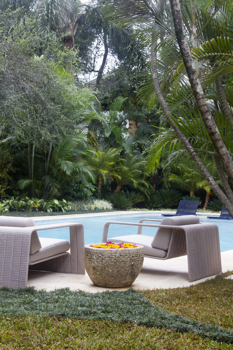 Projeto paisagístico tropical sofisticado em área de piscina, com densa massa vegetal composta por palmeiras de diferentes portes, arbustos tropicais e forração de grama-preta criando camadas de texturas e volumes que garantem total privacidade e imersão na natureza. O mobiliário contemporâneo em fibra sintética cinza com almofadas claras e o vaso de pedra esculpida com flores flutuantes conferem elegância e refinamento ao espaço de contemplação junto à piscina de bordas suaves revestida em pedr, estilo Moderno, Tropical, elementos Piscina retangular com bordas arredondadas em pedra natural clara, Par de poltronas em fibra sintética trançada cinza com almofadas brancas, Vaso de pedra talhada com flores flutuantes amarelas e roxas, Espreguiçadeiras azuis ao fundo da piscina, Piso de pedra natural tipo mármore travertino ao redor da piscina, Maciço vegetal tropical denso como pano de fundo, plantas Palmeira-real, Palmeira-areca, Cica, Helicônia, Filodendro, Grama-preta, Grama-esmeralda, Samambaia, Palmeira-rápis, Licuala, Clúsia, Singônio, Agave, Alpínia, Pau-brasil, ambiente relaxante, sofisticado, acolhedor, localização piscina, jardim, área grande, estruturas nenhum, cores Verde em múltiplos tons, Azul da piscina, Cinza claro do mobiliário, Branco das almofadas, Bege do piso em pedra, Amarelo e roxo das flores no vaso, Marrom dos troncos das palmeiras, piso grama, pedra, privacidade cerca_viva, muro_verde, água piscina