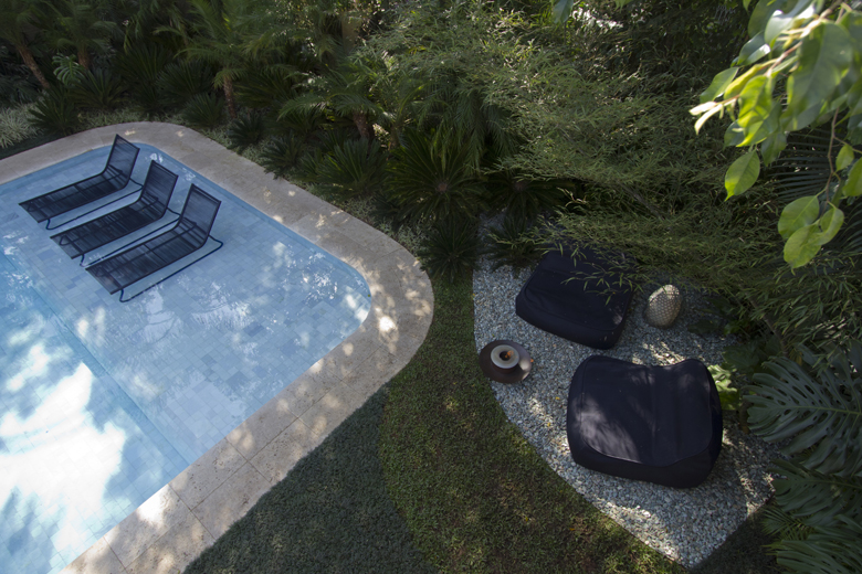 Projeto paisagístico tropical contemporâneo visto de cima, com piscina de formato orgânico com borda arredondada em pedra calcária natural, emoldurada por densa vegetação tropical que cria um verdadeiro refúgio privativo. O lounge externo sobre brita cinza com poltronas escuras contrasta elegantemente com o verde exuberante de palmeiras-leque, bambuzais, filodendros e heliconias que envolvem todo o espaço, proporcionando sombreamento natural e total privacidade. A composição equilibra sofisticaç, estilo Moderno, Tropical, elementos Piscina com formato orgânico e borda em pedra natural calcária, Duas espreguiçadeiras de estrutura metálica preta sobre shelf da piscina, Duas poltronas lounge em tecido náutico preto sobre área de brita, Mesa lateral redonda em madeira ou metal, Vaso decorativo dourado/metalizado tipo lanterna, Área de brita cinza delimitando lounge externo, Bordadura em pedra natural ao redor da piscina, plantas Palmeira-leque, Bambu, Filodendro-guaimbê, Palmeira-areca, Grama-esmeralda, Costela-de-adão, Helicônia, Palmeira-rápis, ambiente relaxante, sofisticado, moderno, acolhedor, localização piscina, jardim, quintal, área medio, estruturas iluminacao, cores verde-escuro, verde-claro, azul-piscina, preto, cinza, bege, dourado, piso grama, brita, pedra, privacidade cerca_viva, água piscina