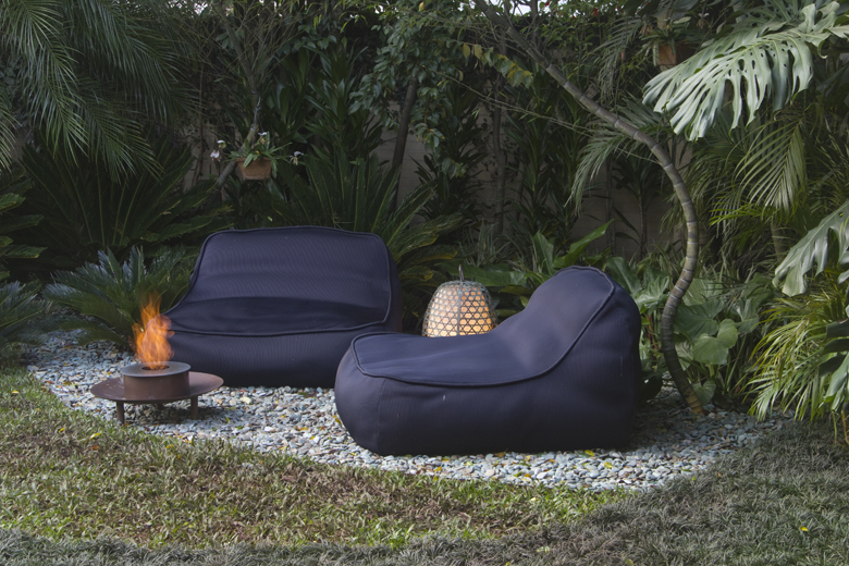 Projeto de jardim tropical contemporâneo que integra uma área de estar intimista emoldurada por densa vegetação de porte médio a grande, criando uma atmosfera de refúgio urbano com forte apelo sensorial. O piso combina gramado esmeralda com bolsão de seixos rolados em tons de branco e cinza, sobre o qual repousam mobiliários em tecido náutico preto de linhas orgânicas, uma lareira ecológica portátil e uma luminária artesanal em tramado de bambu que remete à estética tropical brasileira. A massa , estilo Moderno, Tropical, elementos Lareira ecológica portátil sobre base metálica circular, Poltrona tipo bean bag em tecido náutico preto, Sofá tipo bean bag dois lugares em tecido náutico preto, Luminária decorativa de piso em formato de pinha com tramado em bambu/rattan, Forração de seixos brancos e cinzas delimitando a área de estar, Gramado natural circundando a área de pedriscos, plantas Palmeira-rabo-de-peixe, Costela-de-adão, Helicônia, Palmeira-areca, Filodendro, Samambaia, Singônio, Pacová, Palmeira-real, Lírio-da-paz, Grama-esmeralda, ambiente relaxante, moderno, acolhedor, sofisticado, localização jardim, quintal, área medio, estruturas iluminacao, cores verde-escuro, verde-claro, preto, branco, cinza, dourado, laranja-fogo, marrom, piso grama, brita, privacidade cerca_viva, muro_verde, água nenhum