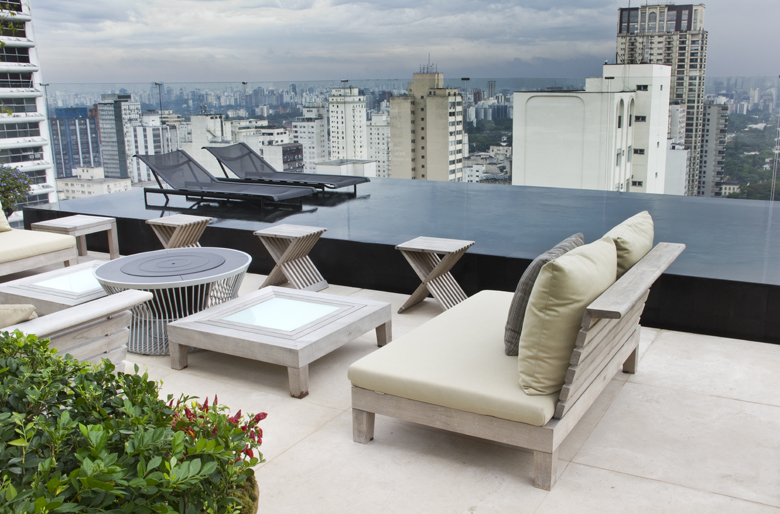 Terraço panorâmico de cobertura em edifício de alto padrão em São Paulo, com piscina de borda infinita em revestimento escuro que cria um espelho d'água dramático contra o skyline urbano. O mobiliário combina madeira de demolição envelhecida naturalmente com peças metálicas contemporâneas, formando um lounge sofisticado com daybed estofado em tons neutros e almofadas verde-oliva que dialogam com a vegetação compacta em jardineiras junto ao piso. O paisagismo é contido e minimalista, privilegiand, estilo Moderno, Tropical, elementos piscina de borda infinita com revestimento escuro, espreguiçadeiras de tela preta com estrutura metálica, sofá/daybed de madeira maciça com estofado bege e almofadas verde-oliva, mesa de centro baixa de madeira com tampo de vidro embutido, banquetas laterais de madeira com design X dobrável, mesa lateral redonda com estrutura ripada metálica e tampo com queimador embutido (fire pit), jardineira com vegetação baixa e compacta junto ao piso, piso de porcelanato claro em formato de grandes placas, plantas Buxinho, Clúsia, Pingo-de-ouro, ambiente sofisticado, moderno, relaxante, localização piscina, varanda, área grande, estruturas banco, iluminacao, cores bege, cinza, preto, verde-oliva, branco, verde-escuro, piso porcelanato, privacidade nenhum, água piscina