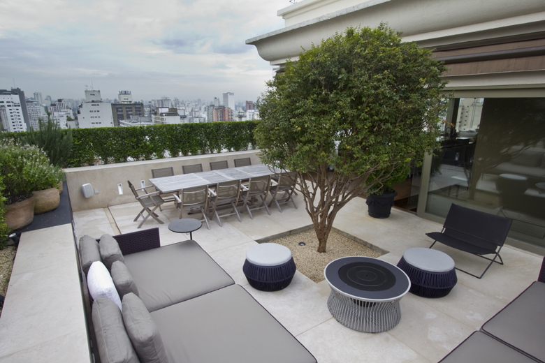 Terraço de cobertura em estilo contemporâneo sofisticado localizado em São Paulo, com uma pitangueira adulta como ponto focal central plantada em área de pedrisco, cercada por mobiliário de design com linhas limpas em tons neutros. A cerca viva de buxinho cuidadosamente podada em toda a extensão do parapeito proporciona privacidade e emoldura a vista panorâmica da cidade, enquanto vasos de cerâmica com aromáticas como alecrim e lavanda acrescentam textura e perfume ao espaço. O projeto equilibra, estilo Moderno, Classico, elementos sofá modular cinza com almofadas brancas, mesa de jantar retangular com tampo de mármore e cadeiras dobráveis de madeira, pufes redondos em tom roxo/marinho, mesa de centro circular com base metálica tipo turbina, poltrona preta de design contemporâneo com estrutura metálica, mesa lateral redonda escura, vasos de cerâmica rústica em tons terrosos, vaso preto grande para árvore, cerca viva podada como mureta verde perimetral, piso de porcelanato bege de grande formato, área de brita/pedrisco ao redor da árvore central, guarda-corpo com acabamento branco clássico, porta de correr em vidro com esquadria escura, plantas Pitangueira, Buxinho, Alecrim, Lavanda, ambiente sofisticado, moderno, relaxante, localização varanda, área medio, estruturas banco, iluminacao, cores cinza, branco, bege, marinho, preto, verde, terracota, piso porcelanato, brita, privacidade cerca_viva, água nenhum
