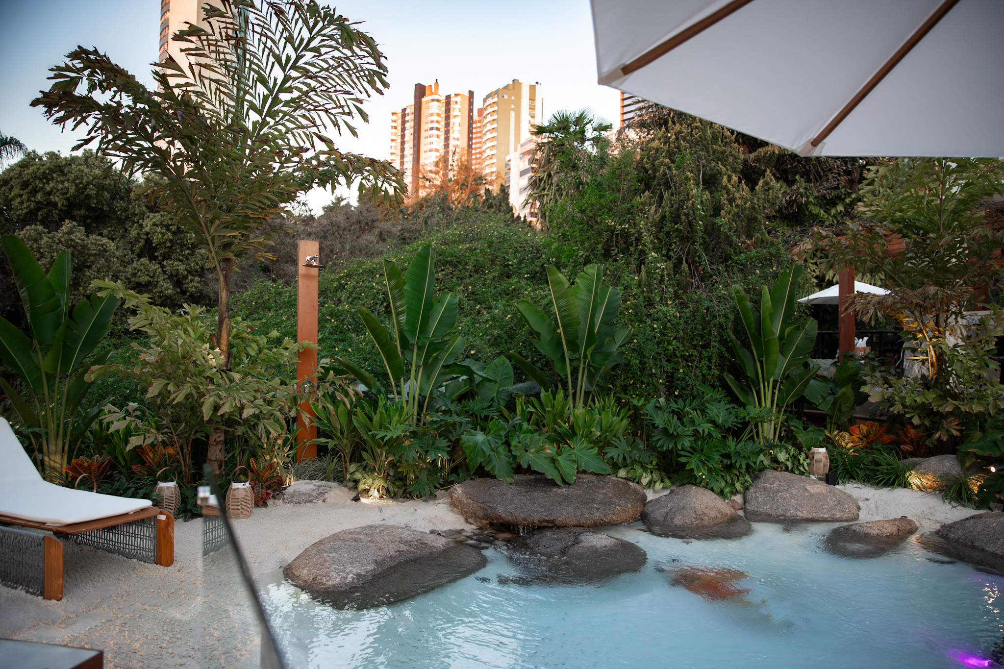 Projeto paisagístico de alto padrão em área urbana, com piscina naturalística de formato orgânico emoldurada por rochas brutas e faixa de areia, criando a atmosfera de uma lagoa tropical privativa. A vegetação exuberante composta por estrelítzias, filodendros, bananeiras-ornamentais, bromélias e trepadeiras forma um denso cinturão verde que garante total privacidade mesmo com edifícios residenciais ao fundo, enquanto uma ducha em aço corten, iluminação de balizadores e um gazebo complementam a a, estilo Tropical, Moderno, elementos Piscina naturalística com formato orgânico e borda de pedras naturais brutas, Faixa de areia clara ao redor da piscina simulando praia natural, Ducha externa em coluna de aço corten, Espreguiçadeira em estrutura de alumínio com estofado branco, Ombrelone branco com estrutura em madeira, Lanternas decorativas de mesa em estilo rústico, Iluminação de jardim embutida no solo tipo balizador LED, Rochas naturais de grande porte integradas à piscina, Seixos rolados no interior da piscina, Gazebo ao fundo com cobertura escura, Muro verde formado por trepadeiras densas, plantas Helicônia, Bananeira-ornamental, Ave-do-paraíso / Estrelítzia-branca, Filodendro-guaimbê, Costela-de-adão, Bromélia, Palmeira-fênix, Jabuticabeira ou árvore de porte médio nativa, Unha-de-gato, Singônio, Cróton, Antúrio, Samambaia, Pata-de-elefante, ambiente relaxante, sofisticado, acolhedor, localização piscina, jardim, quintal, área grande, estruturas gazebo, iluminacao, cores Verde intenso, Verde-escuro, Areia / bege claro, Marrom aço corten, Cinza das pedras, Azul-claro (água da piscina), Branco (ombrelone e espreguiçadeira), Vermelho / vinho (bromélias e crótons), Dourado (luz do entardecer), piso pedra, brita, privacidade cerca_viva, muro_verde, água piscina