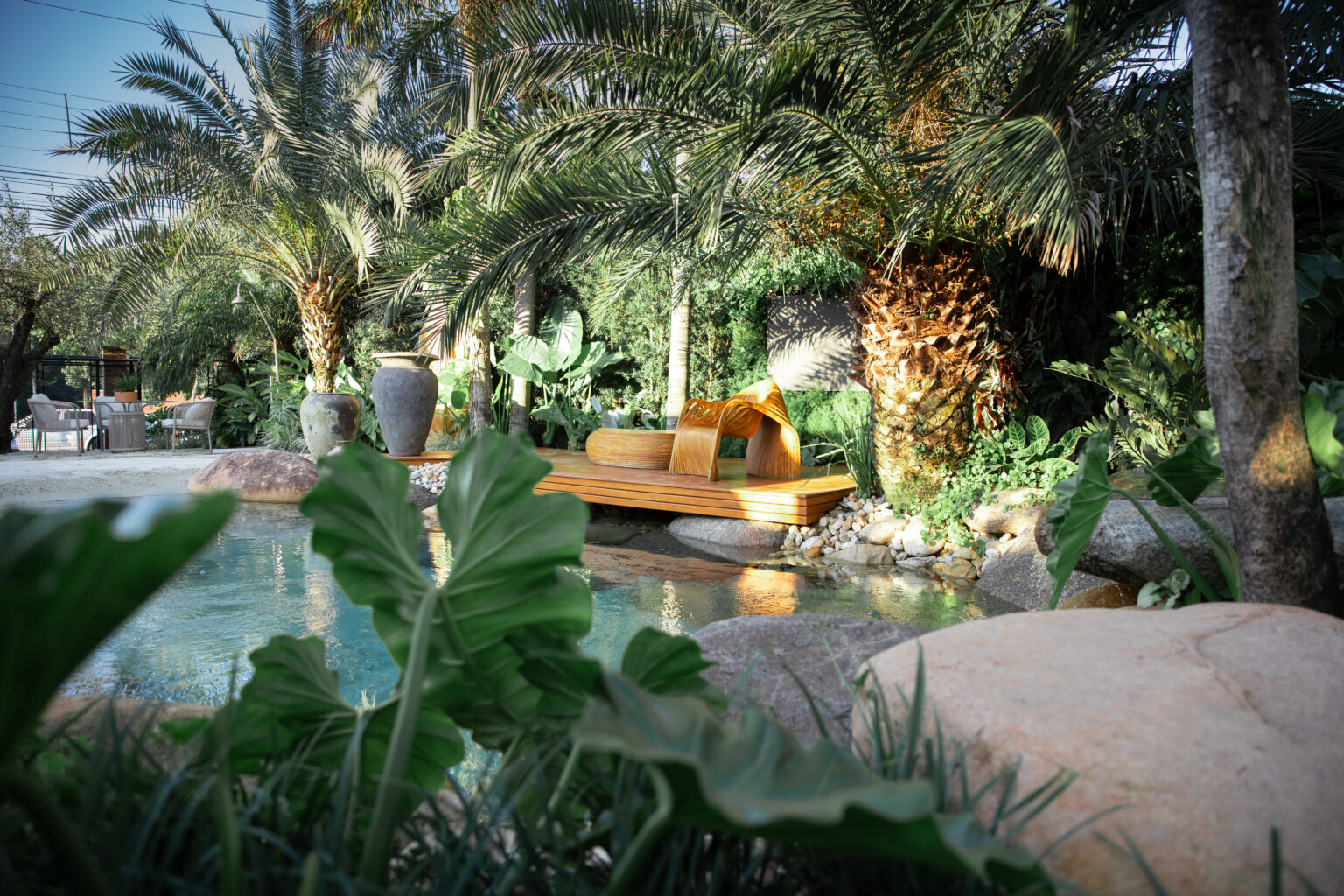 Projeto paisagístico tropical sofisticado de grande porte que integra uma piscina de desenho naturalístico com bordas em pedra bruta a um exuberante jardim de palmeiras-tamareiras, filodendros e alocásias, criando uma atmosfera de oásis tropical privativo. Um elegante deck de madeira nobre se projeta sobre a água, destacando um banco escultural em madeira curvada de linhas orgânicas contemporâneas, ladeado por vasos rústicos de cerâmica e iluminação cênica nos troncos das palmeiras. A composição, estilo Tropical, Moderno, elementos Piscina naturalística com bordas orgânicas em pedra natural, Deck de madeira nobre elevado sobre a água, Banco escultural em madeira curvada com design orgânico contemporâneo, Vasos de cerâmica rústica de grande porte em tons de cinza, Pedras roladas de rio de grande porte como elementos decorativos, Seixos brancos e pedriscos decorativos na composição do paisagismo, Iluminação embutida direcionada nos troncos das palmeiras, Área de estar ao fundo com mobiliário em fibra sintética, plantas Palmeira-tamareira, Palmeira-real, Filodendro-guaimbê, Costela-de-adão, Taioba-ornamental, Bananeira-ornamental, Helicônia, Estrelítzia-branca, Pleomele, Oliveira, Singônio, Grama-preta, Peperômia, ambiente relaxante, sofisticado, acolhedor, localização piscina, jardim, quintal, área grande, estruturas banco, iluminacao, cores verde-escuro, verde-claro, marrom-dourado, bege, cinza-pedra, turquesa-água, branco-seixo, mel, piso deck, pedra, brita, privacidade cerca_viva, água piscina