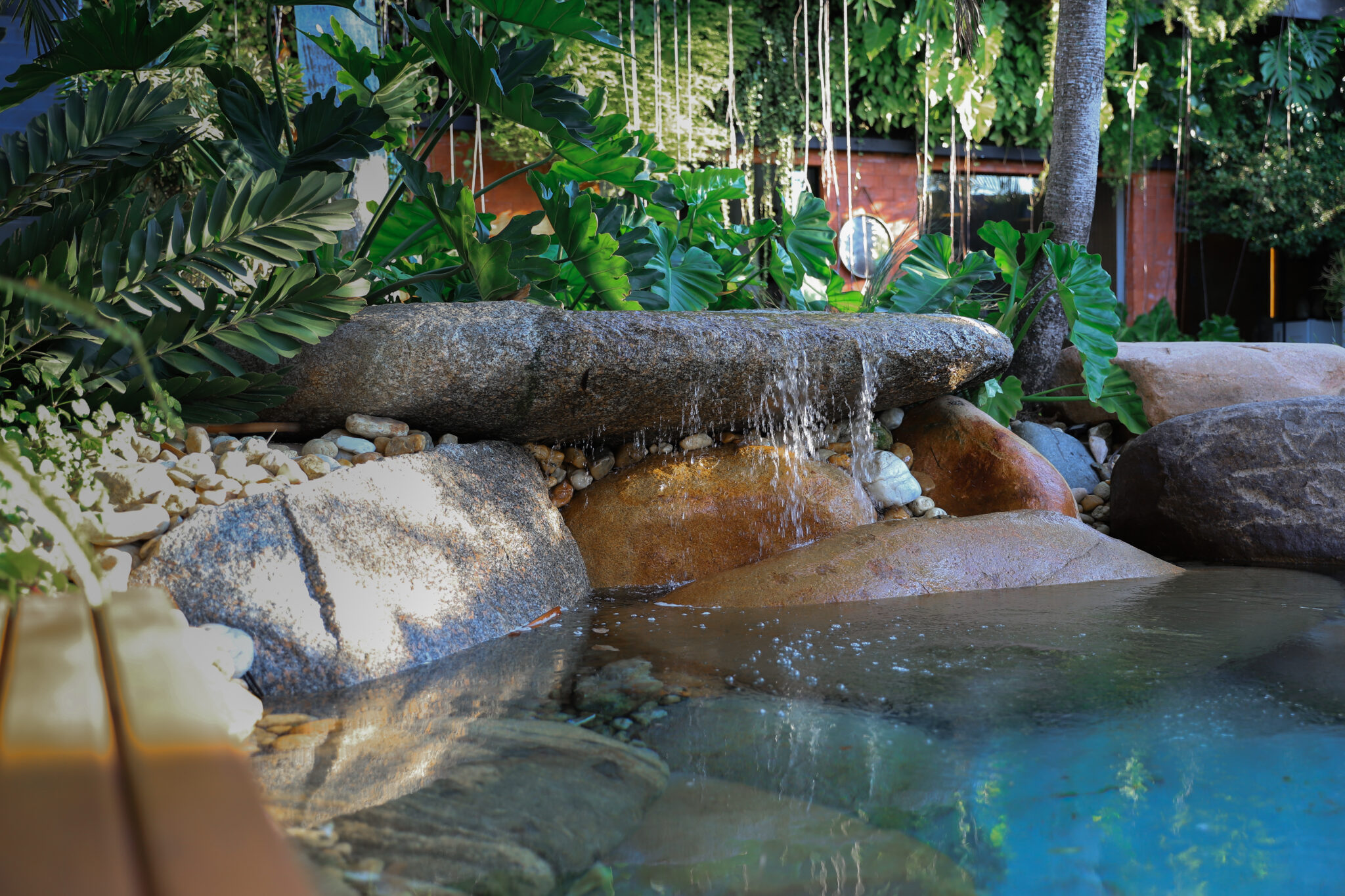 Projeto de piscina naturalista com design biofílico que integra grandes rochas graníticas brutas formando uma cascata com queda d'água sobre um espelho d'água cristalino, evocando uma gruta tropical. A vegetação exuberante de espécies tropicais como costelas-de-adão e filodendros, combinada com plantas pendentes que descem do alto, cria um envolvimento selvagem e imersivo ao redor da estrutura rochosa. O deck de madeira ripada no acesso e os seixos rolados entre as pedras complementam a atmosfer, estilo Tropical, Moderno, elementos Cascata natural em pedra granítica com queda d'água sobre rochas, Pedras graníticas brutas de grande porte compondo o bordo da piscina naturalista, Seixos rolados brancos e bege como acabamento entre as rochas, Piscina naturalista com água cristalina em tom azul-esverdeado, Pedra horizontal posicionada como ponte/vertedouro criando a cascata, Deck em madeira ripada na borda inferior esquerda, Muro de tijolos aparentes ao fundo parcialmente encoberto pela vegetação, Tronco de palmeira integrado à composição rochosa, Pequena escultura decorativa de tartaruga sobre as pedras, plantas Costela-de-adão, Filodendro, Palmeira, Cipó-chumbo / planta pendente com raízes aéreas, ambiente relaxante, sofisticado, acolhedor, localização piscina, jardim, quintal, área medio, estruturas iluminacao, cores verde-escuro, verde-tropical, cinza-granito, bege, marrom-terroso, azul-cristalino, branco, terracota, piso pedra, deck, brita, privacidade muro_verde, água piscina, fonte