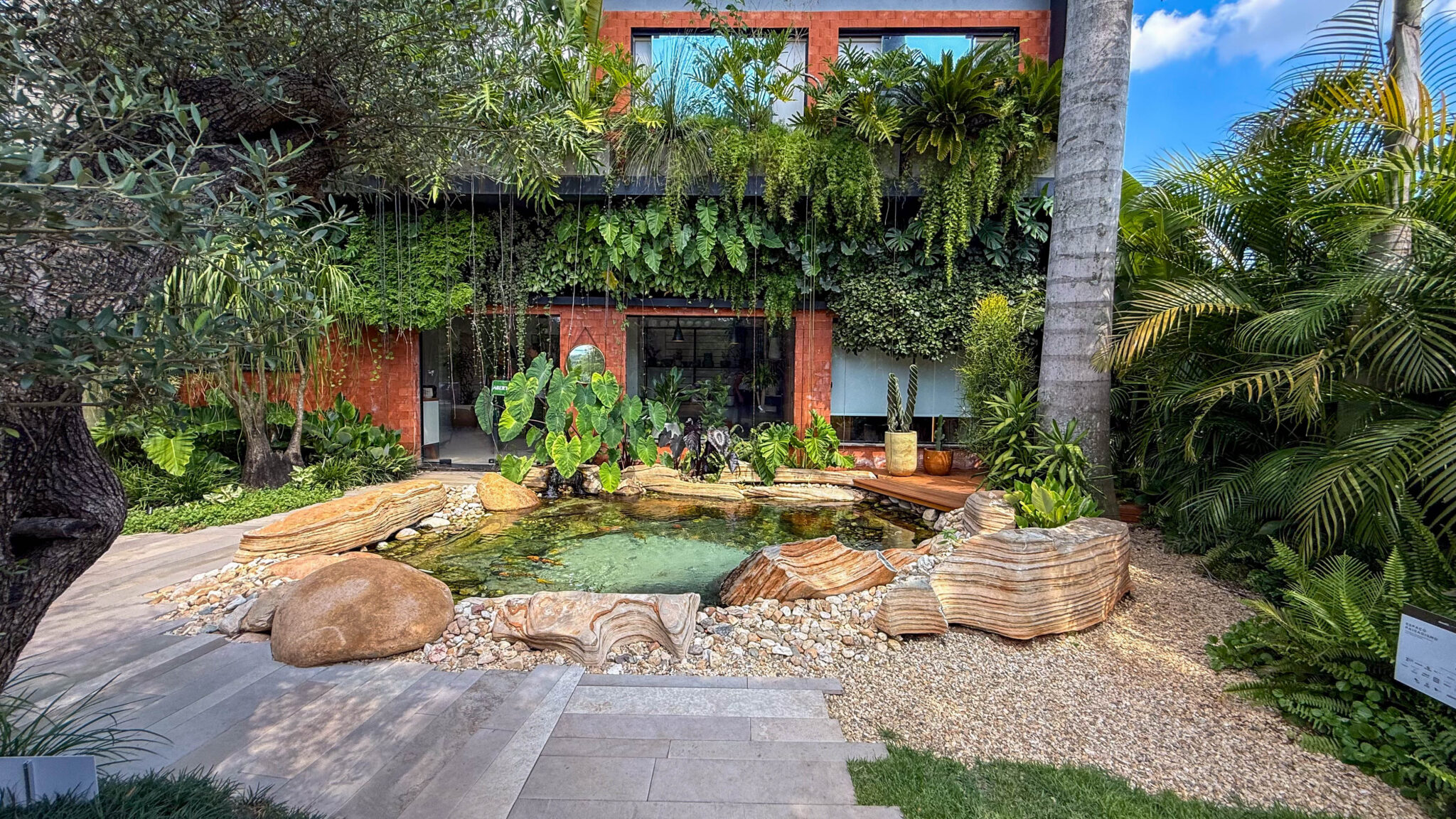 Projeto paisagístico tropical contemporâneo de alto padrão que integra um exuberante jardim vertical na fachada de tijolos aparentes com um lago ornamental central emoldurado por imponentes rochas de arenito estratificado e seixos rolados. A composição mescla espécies tropicais de grande porte como oliveira centenária, palmeiras-reais e alocásias com forrações de samambaias e jiboias pendentes que criam cortinas verdes vivas com fios d'água em cascata. O resultado é um ambiente de contemplação s, estilo Moderno, Tropical, elementos Lago ornamental com água cristalina esverdeada e possíveis carpas, Rochas ornamentais de arenito com veios estratificados, Pedra boulder arredondada de granito, Seixos rolados de rio em tons creme e bege, Piso de porcelanato retangular em tons de cinza, Jardim vertical com múltiplas espécies na fachada, Deck de madeira cumaru junto ao lago, Vasos cerâmicos decorativos em tons terrosos, Cascata sutil com fios de água caindo do jardim vertical superior, Brita decorativa em granulometria fina tom areia, Fachada em tijolos aparentes cor terracota, Esquadrias em alumínio preto com vidro, Grama esmeralda (Zoysia japonica) em bordadura, plantas Oliveira, Costela-de-adão, Taioba-gigante, Filodendro, Samambaia, Hera, Palmeira-real, Palmeira-areca, Renda-portuguesa, Barba-de-serpente, Asplenium, Singônio, Columeia, Lambari, Cactus-coluna, Jiboia, Peperômia, Antúrio, Palmeira-rabo-de-peixe, ambiente sofisticado, relaxante, moderno, localização jardim, fachada, entrada, área grande, estruturas iluminacao, cores verde-escuro, verde-claro, terracota, bege, areia, cinza, marrom, creme, esverdeado-agua, piso porcelanato, brita, pedra, grama, deck, privacidade muro_verde, cerca_viva, água espelho_dagua, fonte