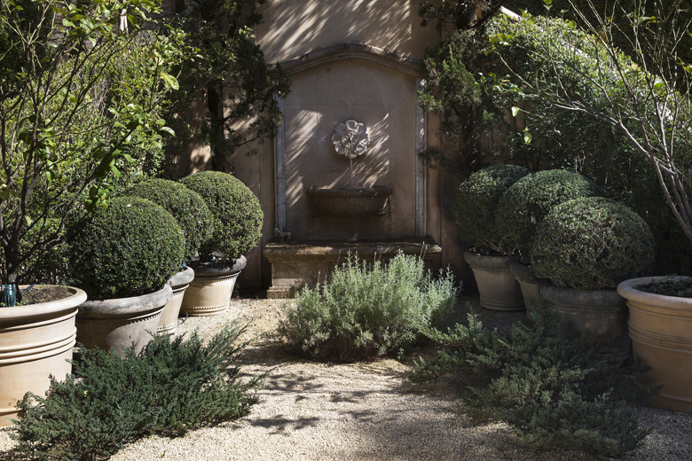 Jardim de inspiração provençal clássica estruturado em torno de uma elegante fonte de parede em pedra calcária com medalha escultórica, ladeada por topiarias esféricas de buxinho em vasos de terracota dispostos simetricamente sobre piso de pedrisco. Herbáceas mediterrâneas como alecrim e lavanda preenchem o primeiro plano com texturas aromáticas prateadas, enquanto trepadeiras como hera e jasmim-estrela emolduram organicamente a estrutura arquitetônica do muro, criando um recanto de intimidade e, estilo Classico, elementos Fonte de parede em pedra calcária com medalha escultórica ornamental, Vasos de barro e terracota em formato cônico de grande porte, Topiaria esférica de buxinho em vasos alinhados simetricamente, Muro de alvenaria com acabamento em reboco envelhecido e moldura clássica, Bacia receptora de água em pedra lavrada sob a bica da fonte, plantas Buxinho, Alecrim, Lavanda, Hera, Jasmim-estrela, ambiente sofisticado, relaxante, acolhedor, localização jardim, muro, entrada, área medio, estruturas iluminacao, nenhum, cores verde-escuro, verde-acinzentado, bege, areia, cinza-pedra, terracota, piso brita, privacidade cerca_viva, muro_verde, água fonte
