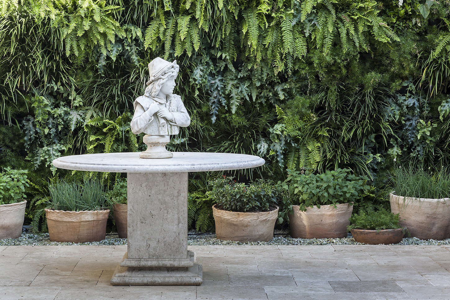 Projeto de paisagismo que combina a exuberância tropical de um jardim vertical repleto de samambaias, aspargos-pendentes e trepadeiras com a elegância clássica europeia de uma escultura em busto de mármore branco sobre mesa de pedra travertino. A composição utiliza vasos artesanais de terracota com ervas aromáticas e folhagens dispostos ao longo da base do muro verde, criando uma transição harmoniosa entre o piso de pedra natural e a parede vegetal. O resultado é um ambiente sofisticado de inspi, estilo Classico, Tropical, elementos Mesa redonda de mármore travertino com base retangular em pedestal, Busto escultórico em mármore branco de figura feminina com chapéu estilo neoclássico, Jardim vertical cobrindo integralmente o muro de fundo, Vasos artesanais de barro/terracota em diferentes tamanhos alinhados na base do muro verde, Piso em placas de pedra natural tipo São Tomé ou calcário em formato retangular, Faixa de brita/seixos cinza entre o piso e os vasos de barro, plantas Samambaia-de-boston, Samambaia-americana, Renda-portuguesa, Hera, Lambari, Barba-de-serpente, Cebolinha, Hortelã, Manjericão, Peperômia, Aspargo-pendente, ambiente sofisticado, acolhedor, relaxante, localização jardim, muro, área medio, estruturas nenhum, cores verde-escuro, verde-claro, branco, bege, terracota, cinza, piso pedra, brita, privacidade muro_verde, água nenhum