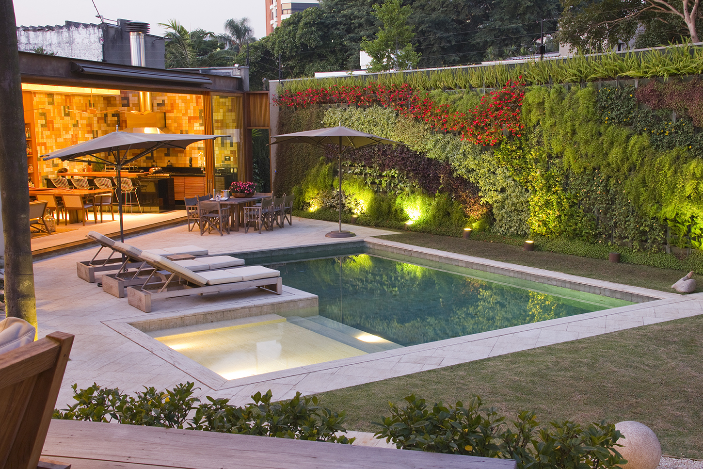 Projeto paisagístico contemporâneo de alto padrão em residência urbana, destacando-se um imponente jardim vertical que reveste todo o muro lateral com composição exuberante de espécies tropicais em tons de verde, roxo e vermelho, iluminado por spots de piso que criam um efeito cênico noturno sofisticado. A piscina retangular de borda infinita em pedra travertino clara integra-se a um spa elevado com iluminação subaquática, ladeada por espreguiçadeiras minimalistas e área de estar sob ombrelones,, estilo Moderno, Tropical, elementos Piscina retangular com borda em pedra natural clara, Spa/hidromassagem integrado à piscina com iluminação subaquática, Jardim vertical de grande porte cobrindo muro divisório, Espreguiçadeiras de madeira com estofamento branco, Mesa de jantar externa com cadeiras sob ombrelones cinza, Cozinha gourmet integrada com abertura total em vidro e painel dourado/amadeirado, Banquetas altas tipo bar no balcão interno, Iluminação cênica de piso (spots uplight) junto ao muro verde, Balizadores de jardim ao longo do gramado, Gramado bem aparado entre piscina e muro verde, Piso em mármore travertino no deck da piscina e áreas de circulação, Churrasqueira com coifa metálica visível no telhado, plantas Bougainvillea, Renda-portuguesa, Aspargo-pluma, Grama-esmeralda, Peperômia, Tradescância-roxa, Samambaia-americana, Dinheiro-em-penca, Clúsia, Lírio-da-paz, Columéia, Barba-de-serpente, Tuia-holandesa, Russélia, Singônio, Azaleia, ambiente sofisticado, moderno, relaxante, localização piscina, quintal, muro, área grande, estruturas churrasqueira, iluminacao, cores verde-escuro, verde-claro, vermelho-bougainvillea, roxo, bege-travertino, dourado, cinza, branco, âmbar, piso grama, pedra, privacidade muro_verde, cerca_viva, água piscina, espelho_dagua