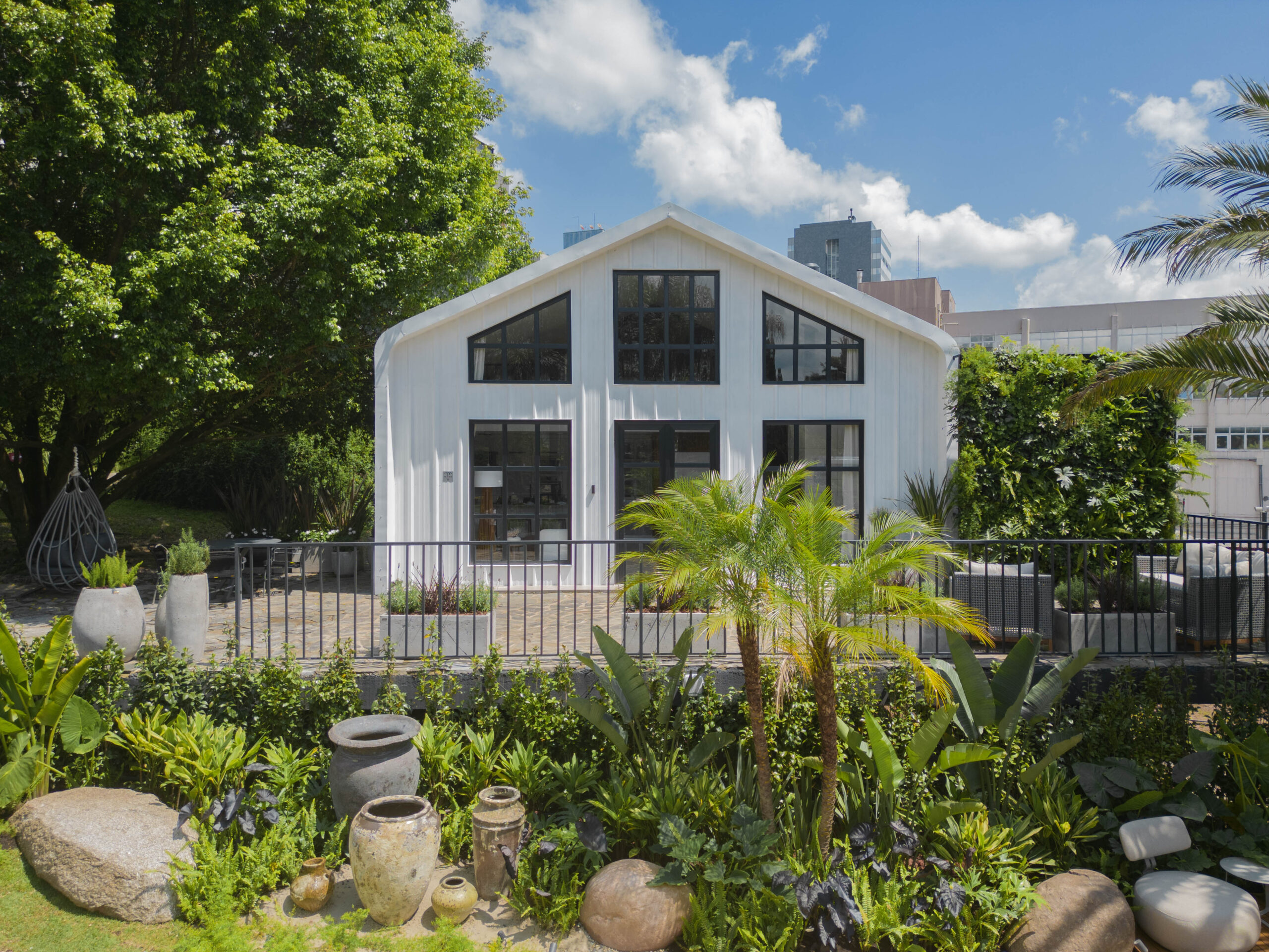 Projeto paisagístico tropical contemporâneo que emoldura uma edificação estilo barn house branca com esquadrias pretas industriais, criando um contraste sofisticado entre a arquitetura moderna e a exuberância da vegetação subtropical. O jardim frontal é composto por camadas de palmeiras, helicônias, estrelítzias e filodendros dispostos em massas verdes densas, intercalados com vasos de concreto escultóricos e cerâmicas rústicas envelhecidas que conferem textura e personalidade ao espaço. Um impo, estilo Moderno, Tropical, elementos Vasos de concreto em formatos orgânicos de diferentes alturas, Vasos de cerâmica rústica envelhecida (estilo wabi-sabi), Pedras naturais grandes tipo matacões decorativos, Grade metálica preta estilo industrial como guarda-corpo, Cadeira suspensa tipo balanço em rattan/fibra sintética, Muro verde / jardim vertical na lateral direita da edificação, Piso em pedra natural tipo paralelepípedo/mosaico português na área de estar, Canteiros elevados com forrações verdes densas, Edificação estilo barn house branca com esquadrias pretas, Floreiras integradas ao paisagismo junto à fachada, plantas Palmeira-areca, Helicônia, Bananeira-ornamental, Ave-do-paraíso / Estrelítzia, Buxinho, Filodendro, Costela-de-adão, Dracena-vermelha, Taioba-ornamental, Coléus, Antúrio-negro, Árvore-da-felicidade ou espécie nativa de grande porte, Palmeira-fênix, Ripsális ou trepadeira em muro verde, Asplenio / Ninho-de-passarinho, Agave ou Bromélia, ambiente relaxante, moderno, sofisticado, acolhedor, localização jardim, fachada, entrada, área grande, estruturas banco, iluminacao, cores Verde em múltiplos tons (do claro tropical ao escuro profundo), Branco (fachada e estrutura), Preto (esquadrias e gradil metálico), Cinza (vasos de concreto e pedras), Bege/terracota (vasos cerâmicos envelhecidos), Vinho/bordô (dracenas e folhagens escuras), Azul (céu refletido no conjunto), piso pedra, brita, privacidade cerca_viva, muro_verde, água nenhum