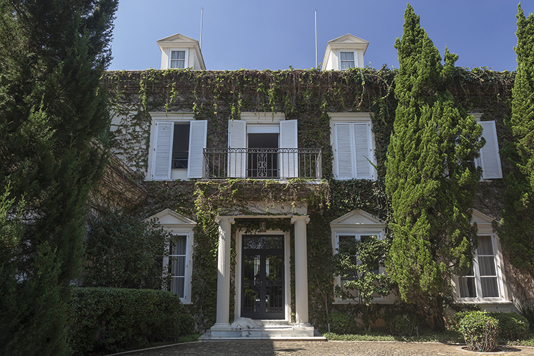 Imponente casarão de estilo neoclássico francês com fachada quase inteiramente coberta por hera e unha-de-gato, criando um dramático muro verde que contrasta elegantemente com as venezianas e molduras brancas das janelas e o gradil de ferro do balcão superior. Dois ciprestes-italianos de grande porte ladeiam a entrada principal com pórtico de colunas dóricas, reforçando a verticalidade e a simetria da composição paisagística de inspiração europeia. A base da fachada é arrematada por topiaria de , estilo Classico, elementos Fachada colonial com colunas neoclássicas brancas, Balcão com gradil de ferro forjado no pavimento superior, Janelas com venezianas brancas em ambos os pavimentos, Mansardas com frontões triangulares no telhado, Pórtico de entrada com frontão triangular e colunas dóricas, Porta principal em vidro e ferro com moldura escura, Muro verde cobrindo quase toda a fachada de alvenaria, Topiaria de buxinho formando cerca viva baixa na base, Piso em paralelepípedo de pedra na entrada, plantas Hera, Cipreste-italiano, Buxinho, Unha-de-gato, Jasmim-estrela, ambiente sofisticado, acolhedor, localização fachada, entrada, muro, área grande, estruturas iluminacao, cores verde-escuro, verde-claro, branco, cinza, marrom, preto, piso pedra, privacidade muro_verde, cerca_viva, água nenhum