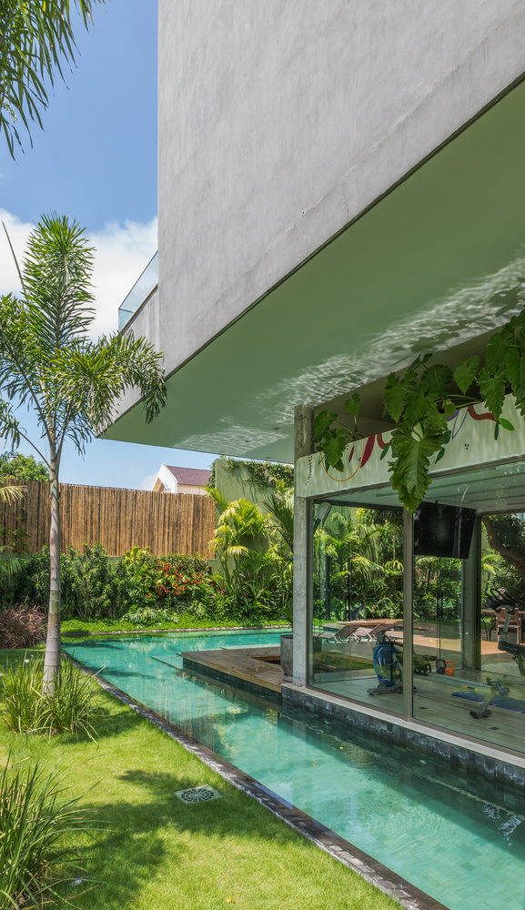 Projeto paisagístico tropical contemporâneo integrado a uma residência de arquitetura brutalista com laje em balanço de concreto aparente e amplos painéis de vidro piso-teto, onde uma piscina em formato de canal linear percorre toda a lateral da edificação, margeada por gramado de esmeralda impecável e canteiros exuberantes de palmeiras, helicônias e alpínias. A vegetação densa e estratificada ao fundo cria uma cortina verde que, aliada à cerca de bambu natural, garante total privacidade ao espa, estilo Moderno, Tropical, elementos Piscina retangular em formato de canal com borda de pedra natural, Deck de madeira sobre a piscina como plataforma de descanso, Cerca de bambu natural como divisa perimetral, Espreguiçadeiras de madeira ao fundo, Piso de vidro/janelas piso-teto com esquadria de alumínio, Laje em balanço de concreto aparente formando cobertura, Pilares de concreto aparente, Ralo circular embutido na borda da piscina, Canteiro tropical volumoso ao fundo, plantas Palmeira-areca, Dracena, Helicônia, Palmeira-real, Bougainvillea, Alpínia, Costela-de-adão, Filodendro, Grama-esmeralda, Palmeira-rápis, Cróton, Jiboia, Pleomele, ambiente relaxante, moderno, sofisticado, localização jardim, piscina, fachada, quintal, área grande, estruturas iluminacao, cores verde-esmeralda, turquesa, branco, cinza-concreto, marrom-madeira, amarelo-bambu, vermelho-floral, piso grama, deck, pedra, privacidade cerca_viva, muro_verde, água piscina