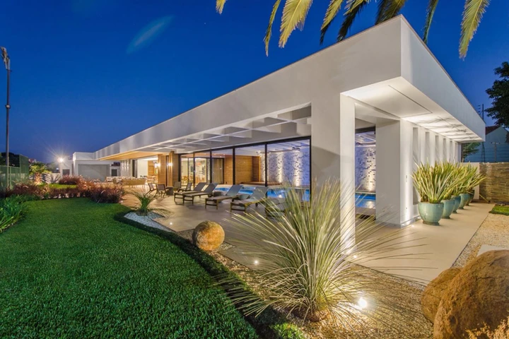 Projeto paisagístico contemporâneo de alto padrão que integra volumetria arquitetônica minimalista com jardim tropical exuberante, utilizando canteiros orgânicos com gramado esmeralda impecável, pedras ornamentais de grande porte e massas vegetais de texturas contrastantes — capins ornamentais, dracenas e palmeiras-fênix. A iluminação cênica com spots de LED embutidos no piso valoriza cada espécie vegetal e as pedras naturais, criando um efeito dramático noturno que dialoga com a piscina ilumina, estilo Moderno, Tropical, elementos Iluminação embutida no piso tipo spot LED direcionada às plantas, Pedras ornamentais naturais de grande porte em tons ocre, Canteiros com bordas orgânicas delimitando gramado e áreas pavimentadas, Vasos cerâmicos em tom verde-água de grande porte, Espreguiçadeiras em área de deck junto à piscina, Piscina com iluminação interna azul e revestimento em pastilhas, Cobertura em laje plana com vigas aparentes em concreto branco, Fachada com painéis de vidro piso-teto e esquadrias pretas, Parede decorativa com revestimento texturizado e iluminação cênica azulada, Piso em porcelanato claro na área da varanda coberta, plantas Palmeira-fênix, Yucca, Capim-do-texas, Dracena-vermelha, Formio, Agave-dragão, Grama-esmeralda, Moréia, Cróton, Bromélias, Clúsia, ambiente sofisticado, moderno, relaxante, localização jardim, fachada, piscina, varanda, área grande, estruturas iluminacao, pergolado, cores verde-escuro, verde-claro, branco, ocre, azul, bege, vermelho-bordô, cinza, piso grama, porcelanato, pedra, privacidade cerca_viva, água piscina