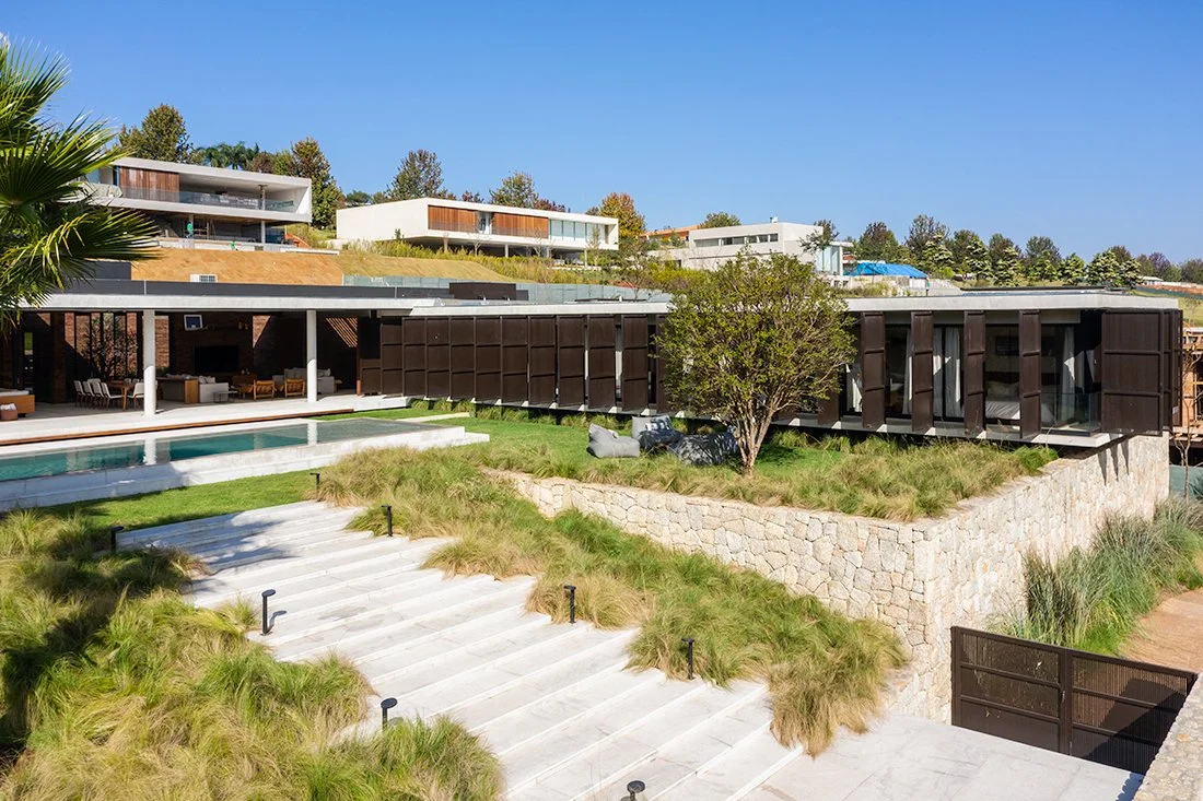 Projeto paisagístico contemporâneo de grande porte integrado a uma residência de arquitetura horizontal com teto verde, brises pivotantes em aço corten e ampla área de lazer com piscina. A escadaria monumental em pedra clara é ladeada por massivos de capim-do-texas (Pennisetum setaceum) que criam movimento e textura orgânica, contrastando com o muro de arrimo em pedra natural rústica e o gramado impecável no platô superior. O paisagismo valoriza a topografia do terreno em desnível, utilizando ve, estilo Moderno, Tropical, elementos Escadaria ampla em pedra natural clara com degraus largos, Muro de arrimo em pedra natural rústica tipo quartzito, Piscina retangular de borda infinita com revestimento claro, Portão de acesso em aço corten ou madeira escura, Balizadores de jardim em LED preto ao longo da escadaria, Cobertura plana com teto verde extensivo, Painéis pivotantes em aço corten como brises nas fachadas, Área gourmet coberta com pilares brancos e estrutura metálica, Pufes e mobiliário de descanso ao ar livre sob a árvore, Pergolado integrado à cobertura da área social, plantas Capim-do-texas, Grama-esmeralda, Palmeira-leque, Jabuticabeira ou árvore ornamental de médio porte, ambiente moderno, sofisticado, relaxante, localização jardim, fachada, piscina, entrada, área grande, estruturas pergolado, churrasqueira, iluminacao, banco, cores verde, bege, marrom-escuro, branco, cinza, dourado-palha, piso grama, pedra, porcelanato, privacidade muro_verde, cerca_viva, água piscina