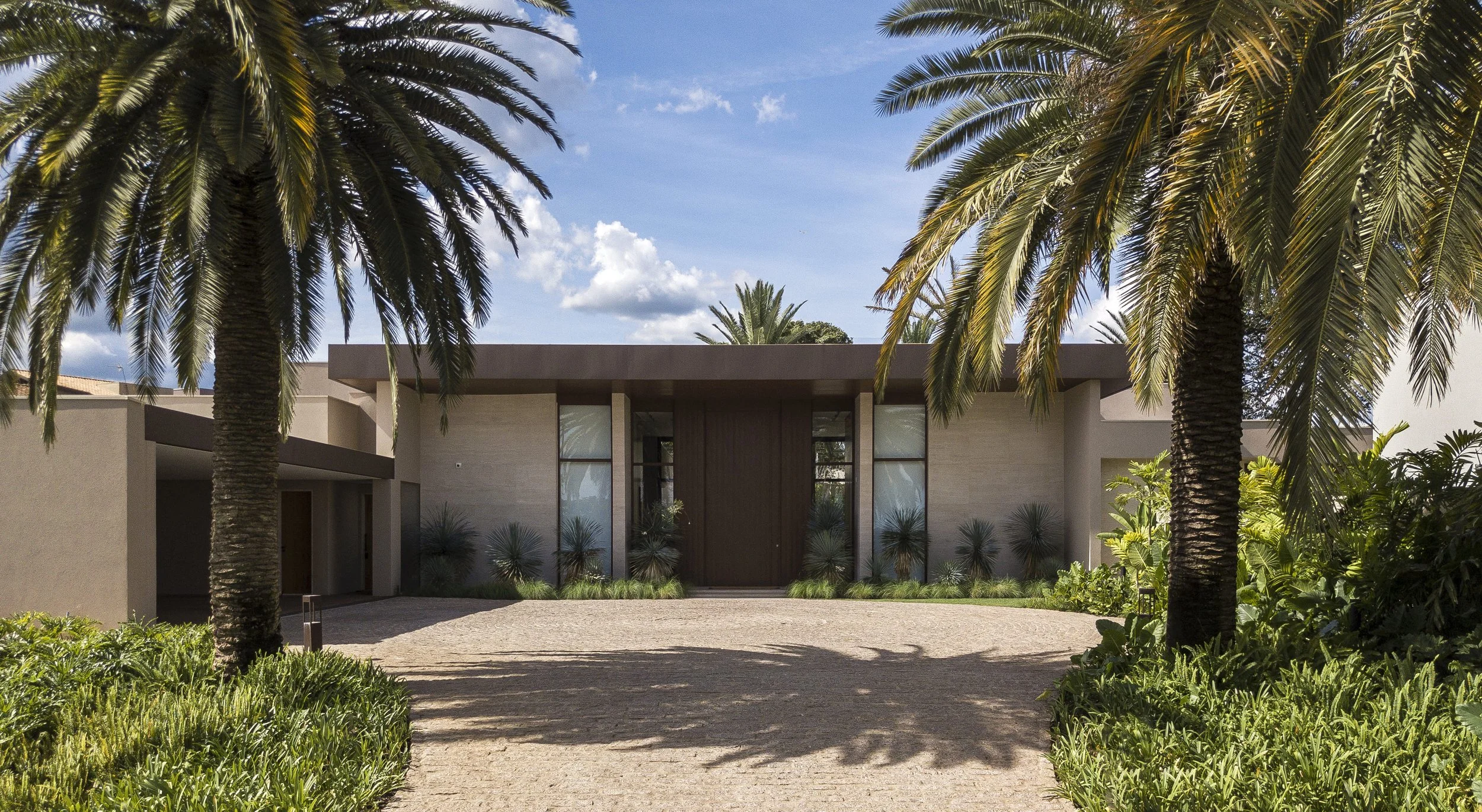 Projeto paisagístico de grande porte em fachada residencial contemporânea, com imponentes palmeiras-das-canárias (Phoenix canariensis) emoldurando simetricamente a entrada e criando um eixo visual majestoso até o portão principal em madeira escura. Canteiros lineares junto à base da fachada combinam agaves, yuccas e dracenas-azuis em composição escultórica de texturas contrastantes sobre forração de liriopes, enquanto massas tropicais de filodendros, helicônias e bananeiras-ornamentais ocupam os, estilo Moderno, Tropical, elementos Piso intertravado em pedra natural tipo paralelepípedo bege, Fachada revestida em mármore travertino ou limestone, Portão principal em madeira maciça escura com pé-direito alto, Grandes panos de vidro temperado emoldurados por pilares de concreto, Platibanda reta em concreto com acabamento em tom marrom/corten, Cobertura em laje plana com projeção em balanço formando marquise, Canteiros lineares junto à fachada com forração e plantas escultóricas, Entrada de veículos lateral com cobertura em laje, Volumetria horizontal com composição de blocos escalonados, plantas Palmeira-das-canárias, Dracena-azul, Yucca, Grama-esmeralda, Helicônia, Bananeira-ornamental, Filodendro, Liriope, Agave-dragão, Moreia, ambiente sofisticado, moderno, localização fachada, entrada, área grande, estruturas iluminacao, cores Bege-areia, Marrom-escuro, Verde-escuro, Verde-azulado, Cinza-claro, Branco, piso pedra, grama, privacidade cerca_viva, água nenhum