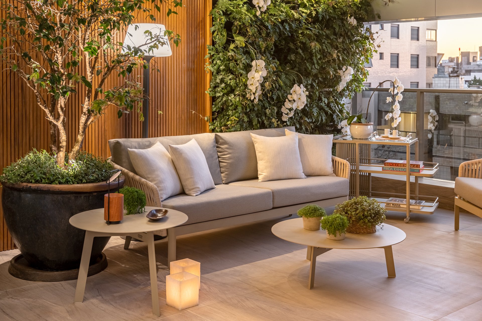 Varanda de apartamento urbano com projeto paisagístico intimista e sofisticado, combinando um painel ripado de madeira natural ao fundo com um exuberante jardim vertical de hera que cria um muro verde, pontuado por orquídeas Phalaenopsis brancas que conferem elegância tropical. O mobiliário contemporâneo em tons neutros — sofá em cordas náuticas, mesas de centro redondas com pés palito e carrinho bar com livros de arte — compõe um ambiente de estar ao ar livre refinado, iluminado por luminária d, estilo Moderno, Tropical, elementos Sofá de três lugares em tecido bege com estrutura em cordas náuticas, Almofadas decorativas em tons de bege e cinza claro, Duas mesas de centro redondas com tampo em tom neutro e pés palito, Vaso de cerâmica escura de grande porte para árvore, Luminária de piso com cúpula branca, Duas luminárias de chão tipo cubos translúcidos com velas/LED, Carrinho bar dourado/metal com livros de arte (Frida Kahlo, Polo, Art House), Vaso cerâmico claro com orquídeas sobre o carrinho, Painel ripado de madeira natural como fundo, Bandeja decorativa sobre carrinho com xícaras, Poltrona em cordas náuticas e madeira parcialmente visível à direita, Cesto de vime com planta verde sobre mesa de centro, Guarda-corpo de vidro da varanda, plantas Orquídea-branca, Hera, Jabuticabeira ou árvore de pequeno porte não identificada com precisão, Musgo-tapete / Selaginela, Baby tears / Lágrima-de-bebê, ambiente relaxante, moderno, acolhedor, sofisticado, localização varanda, área pequeno, estruturas iluminacao, cores bege, cinza claro, verde, branco, madeira natural, dourado, marrom escuro, creme, piso porcelanato, privacidade muro_verde, água nenhum