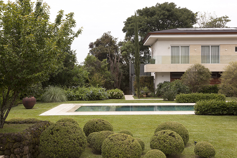 Projeto paisagístico de residência contemporânea de alto padrão que combina a elegância clássica de topiarias esféricas de buxinho em primeiro plano com uma linguagem naturalista nos maciços arbustivos e herbáceos ao redor da piscina retangular em travertino. A composição utiliza camadas vegetais estratificadas — desde o gramado impecável de esmeralda, passando pelos arbustos de podocarpo e clúsia, até ciprestes-italianos e árvores de grande porte que formam um pano de fundo verde exuberante e g, estilo Moderno, Classico, elementos Topiaria esférica de buxinho em massa agrupada no primeiro plano, Piscina retangular com borda em pedra clara tipo travertino, Gramado extenso bem nivelado interligando áreas de plantio, Maciço arbustivo misto ao fundo da piscina com floração branca, Vaso cerâmico decorativo em tom terracota posicionado no canteiro lateral, Fachada residencial contemporânea com revestimento em travertino e vidro, Varanda com guarda-corpo em vidro no pavimento superior, Arborização de grande porte ao fundo criando cortina vegetal de privacidade, plantas Buxinho, Grama-esmeralda, Cipreste-italiano, Hortênsia, Agapanto, Palmeira-imperial, Capim-do-texas, Dracena, Iúca, Jabuticabeira, Podocarpo, Moreia, Clúsia, Viburno, ambiente sofisticado, relaxante, moderno, localização jardim, piscina, fachada, área grande, estruturas iluminacao, cores verde-escuro, verde-claro, branco, bege, terracota, creme, piso grama, pedra, privacidade cerca_viva, água piscina