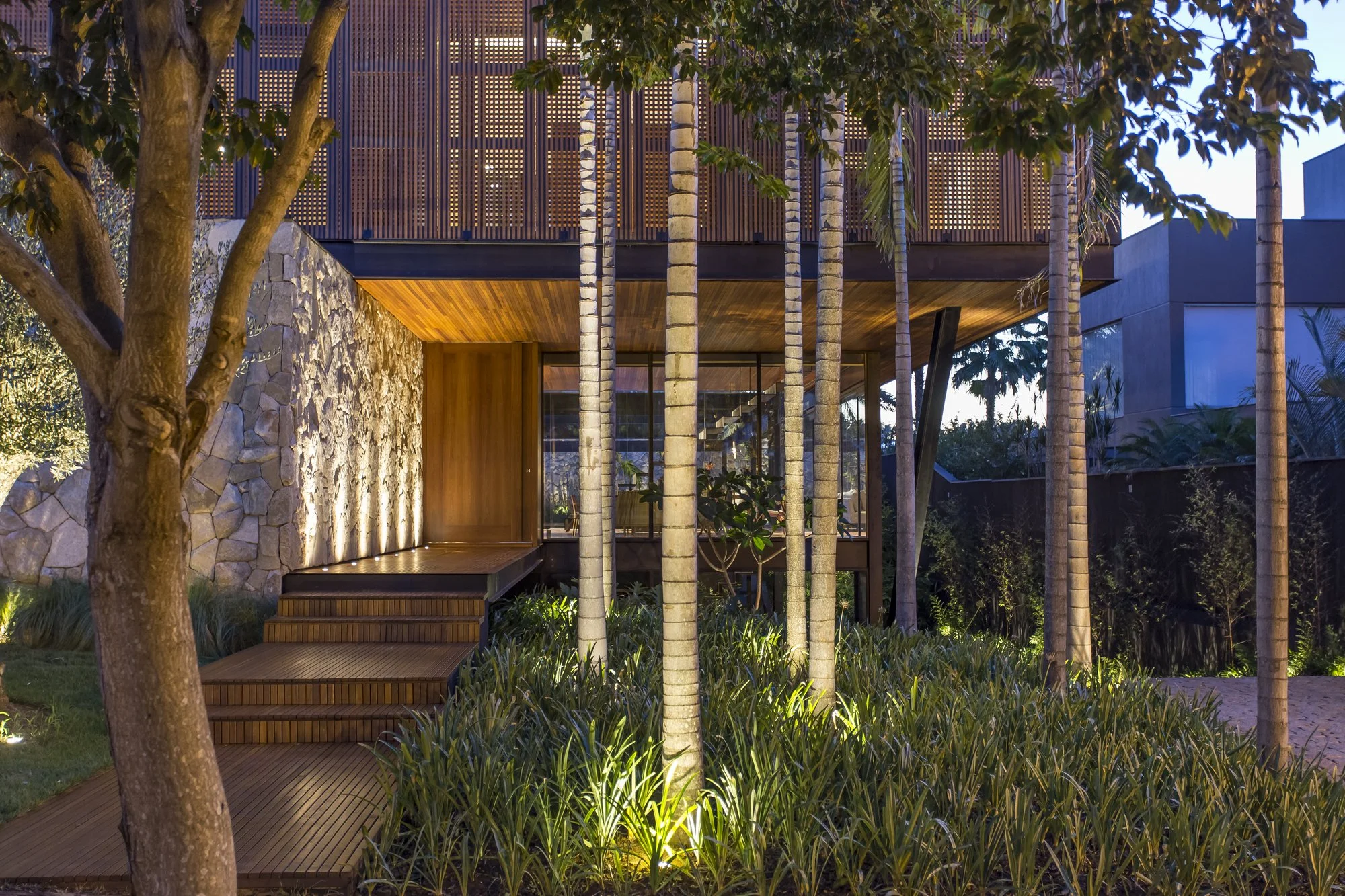 Projeto paisagístico contemporâneo de alto padrão que integra um maciço de palmeiras-reais enfileiradas com forração densa de agapantos e moréias, criando um jardim tropical exuberante diante de uma fachada com muro de pedra natural iluminado por spots ascendentes. O deck de madeira nobre com degraus fluidos conecta o interior da residência ao jardim, enquanto os painéis treliçados (muxarabis) no pavimento superior filtram a luz e conferem privacidade com sofisticação. A iluminação cênica noturn, estilo Moderno, Tropical, elementos Muro de pedra natural irregular com iluminação cênica ascendente, Deck de madeira cumaru com degraus integrados, Forro de madeira no beiral do telhado, Painéis treliçados de madeira com padrão vazado no pavimento superior (muxarabi), Esquadrias de vidro piso-teto com perfil de alumínio preto, Porta pivotante de madeira em tom mel, Iluminação embutida no piso para destaque das palmeiras e muro de pedra, Estrutura de concreto e aço aparente nos pilares, plantas Palmeira-real, Agapanto, Moreia, Oliveira, Barba-de-serpente, Helicônia, Palmeira-açaí, Clúsia, ambiente sofisticado, moderno, acolhedor, relaxante, localização fachada, jardim, entrada, área grande, estruturas iluminacao, cores Verde intenso, Marrom madeira (cumaru/ipê), Cinza pedra natural, Branco dos troncos de palmeira, Dourado da iluminação quente, Azul crepuscular do céu, piso deck, pedra, privacidade cerca_viva, água nenhum
