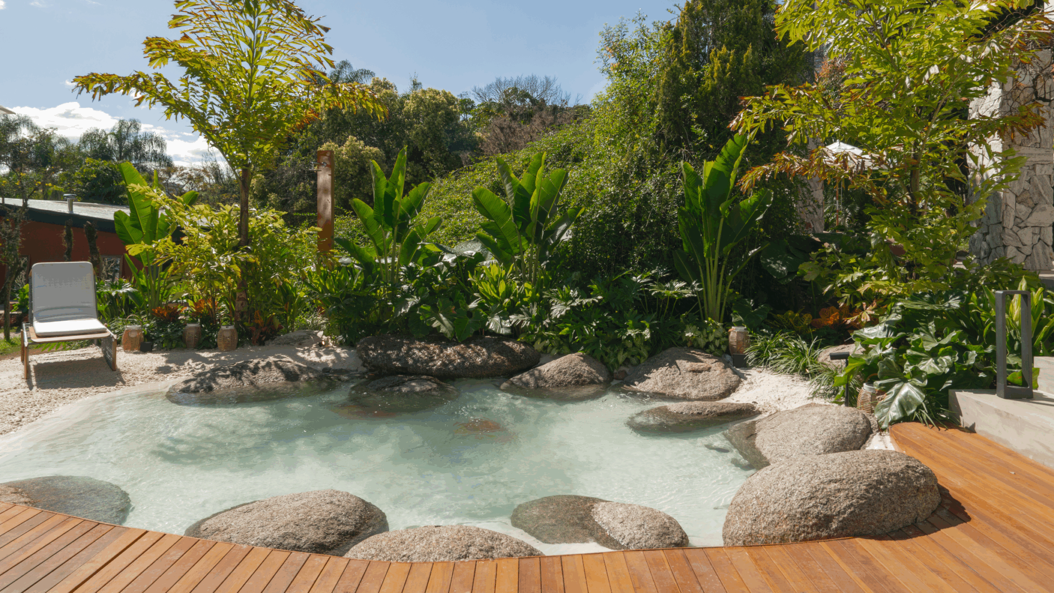 Projeto de paisagismo tropical contemporâneo integrado a uma piscina de formato orgânico que simula uma lagoa natural, com matacões de granito dispostos de forma naturalista tanto dentro quanto nas bordas da água cristalina rasa, circundada por deck de madeira nobre e faixa de areia clara. A vegetação exuberante compõe um maciço denso de espécies tropicais — bananeiras-ornamentais, estrelítzias-gigantes, filodendros e heliconias — criando uma cortina verde envolvente que garante privacidade e am, estilo Tropical, Moderno, elementos Piscina natural com formato orgânico imitando lagoa tropical, Pedras naturais de grande porte (matacões de granito) dentro e ao redor da piscina, Deck em madeira cumaru/ipê com réguas longitudinais, Areia clara compactada simulando praia natural, Espreguiçadeira em alumínio com estofado branco, Luminárias balizadoras em aço escovado junto ao deck, Muro de pedra natural (tipo canjiquinha) na fachada lateral direita, Vasos cerâmicos rústicos decorativos entre as pedras, Guarda-sol/ombrelone branco ao fundo à direita, plantas Helicônia, Bananeira-ornamental, Estrelítzia-gigante / Árvore-do-viajante, Filodendro, Costela-de-adão, Alpínia, Palmeira-areca, Calateia, Aglaonema, Lírio-da-paz, Guaimbê, Jabuticabeira ou árvore de médio porte nativa, Pacová, Taioba-ornamental, ambiente relaxante, sofisticado, acolhedor, localização piscina, jardim, quintal, área medio, estruturas iluminacao, cores verde-escuro, verde-claro, bege-areia, cinza-granito, marrom-madeira, branco, azul-celeste, piso deck, pedra, brita, privacidade cerca_viva, água piscina