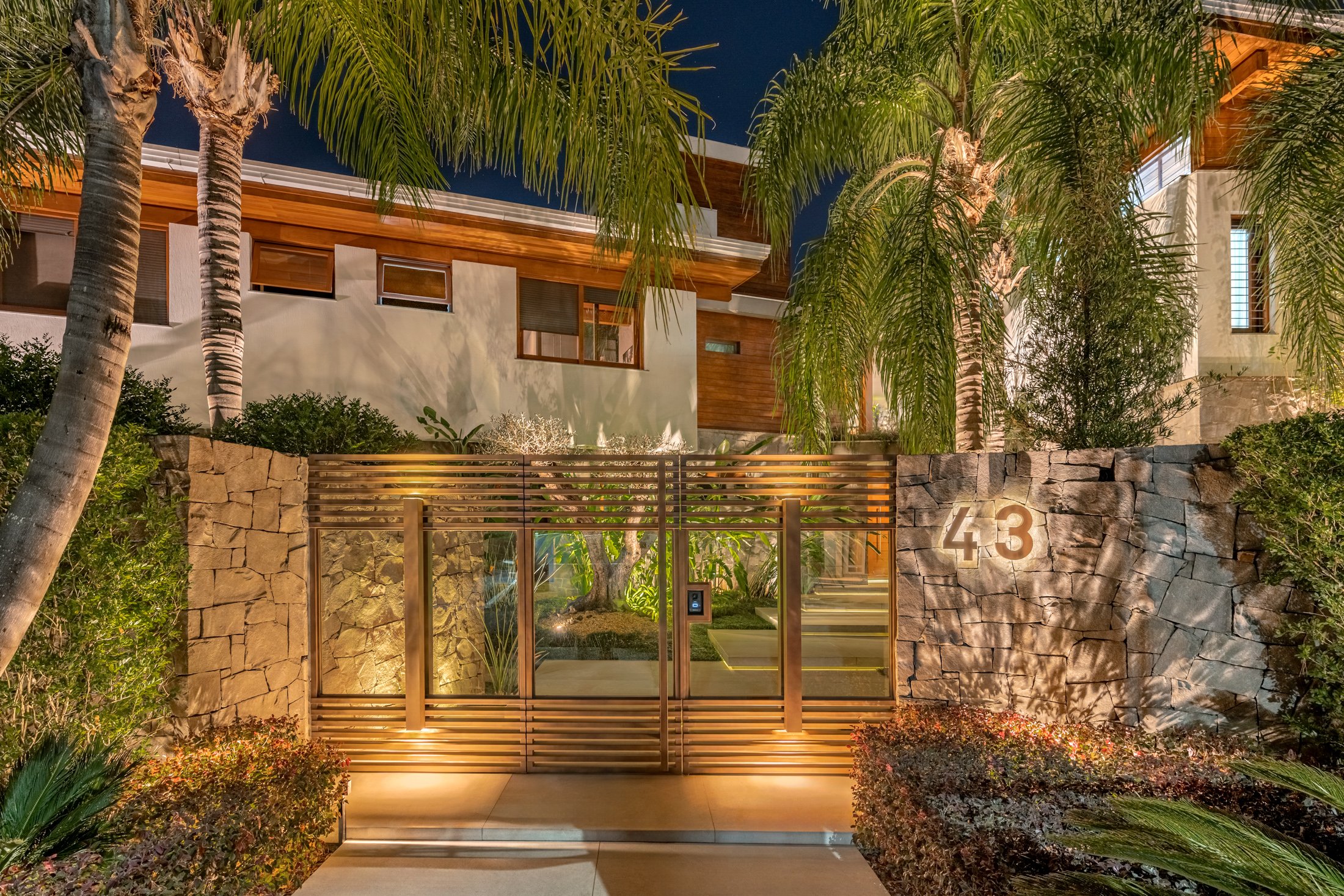 Fachada residencial de alto padrão com linguagem contemporânea tropical, onde o muro de pedra natural rústica se integra harmoniosamente ao portão de alumínio escovado com vidro, criando uma entrada imponente e sofisticada iluminada por spots de piso em tom âmbar quente. Palmeiras-reais e fênix de porte elevado emolduram a composição vertical, enquanto arbustos podados de buxinho e manchas de iresine bordô preenchem os canteiros laterais, proporcionando textura e contraste cromático. O projeto p, estilo Moderno, Tropical, elementos Muro de pedra natural rústica com acabamento irregular, Portão duplo de alumínio escovado com barras horizontais e vidro temperado, Numeração residencial '43' em aço corten fixada no muro de pedra, Iluminação embutida no piso tipo uplighting direcionada às palmeiras e muro, Painel digital de interfone/controle de acesso junto ao portão, Fachada com revestimento em madeira de lei e massa branca, Cobertura com beiral em madeira aparente, Piso de porcelanato bege claro na calçada de acesso, Canteiros elevados laterais com vegetação arbustiva, plantas Palmeira-real, Palmeira-fênix, Jasmim-manga, Buxinho, Iresine, Azaleia, Pingo-de-ouro, Agave, Cróton, ambiente sofisticado, moderno, acolhedor, localização fachada, entrada, muro, área grande, estruturas iluminacao, cores Verde escuro, Verde claro, Dourado, Bege, Cinza pedra, Marrom madeira, Branco, Vermelho bordô, Âmbar (iluminação), piso porcelanato, pedra, privacidade cerca_viva, muro_verde, água nenhum