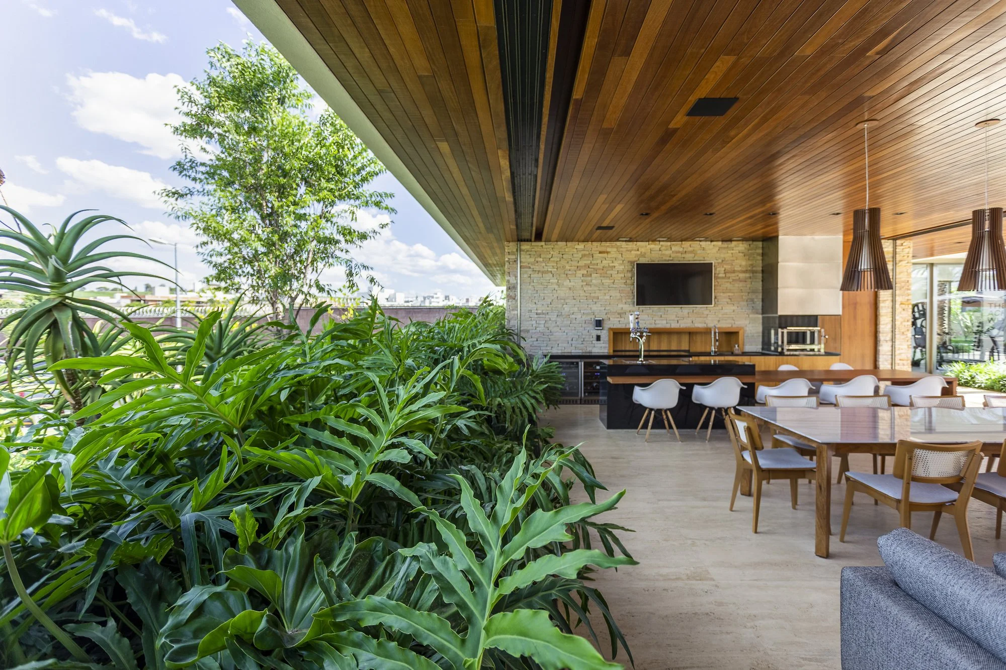 Espaço gourmet contemporâneo de alto padrão com ampla integração interior-exterior, onde um generoso maciço tropical de guaimbês, aloes e pacovás cria uma barreira verde exuberante que delimita a área social coberta do jardim externo, conferindo privacidade e frescor. O forro em madeira nobre ripada se estende do ambiente interno até a projeção da varanda, unificando visualmente a cozinha gourmet com balcão em granito preto, a mesa de jantar para oito pessoas e o canteiro paisagístico tropical. , estilo Moderno, Tropical, elementos forro em madeira ripada natural em tom cumaru, parede revestida em pedra natural canjiquinha, bancada de granito preto com cuba inox, televisão embutida na parede de pedra, mesa de jantar retangular em madeira com tampo de vidro para 8 lugares, cadeiras de jantar em madeira com estofado claro, banquetas brancas estilo Eames no balcão, luminárias pendentes cônicas em madeira escura, sofá cinza parcialmente visível, churrasqueira/área gourmet integrada, piso em porcelanato bege de grande formato, wine cooler/adega embutida sob bancada, forno elétrico embutido, torneira gourmet cromada, plantas Guaimbê, Aloe-candelabro, Helicônia, Pacová, Árvore-da-felicidade, Pau-d'água, ambiente sofisticado, moderno, acolhedor, relaxante, localização varanda, jardim, área grande, estruturas churrasqueira, iluminacao, cores verde intenso, marrom madeira natural, bege areia, preto, branco, cinza claro, tom dourado da madeira do forro, piso porcelanato, privacidade cerca_viva, muro_verde, água nenhum