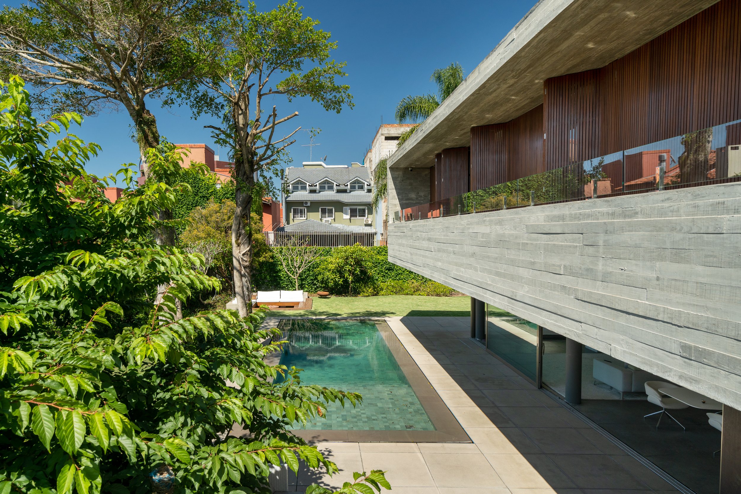 Projeto paisagístico contemporâneo integrado a uma residência brutalista em concreto aparente com generoso balanço estrutural, onde a vegetação tropical exuberante — composta por espécies como jerivá, cafeeiro ornamental e árvores de médio porte — cria uma moldura verde que suaviza a dureza dos materiais e garante privacidade em relação à vizinhança. A piscina retangular de águas cristalinas esverdeadas posiciona-se estrategicamente entre o jardim gramado e o pavimento inferior da casa, estabele, estilo Moderno, Tropical, elementos Piscina retangular de borda infinita com revestimento em tom esverdeado, Parede estrutural em concreto aparente com textura de forma de madeira (board-formed concrete), Daybed branco com estrutura em madeira sobre gramado, Guarda-corpo em vidro temperado na varanda superior, Painéis de brise em madeira cumaru na fachada superior, Piso em placas de porcelanato bege ao redor da piscina, Esquadrias em alumínio preto com vidro amplo no pavimento inferior, Laje em balanço de concreto aparente projetada sobre o jardim, Banqueta alta visível no ambiente interno térreo, plantas Palmeira-jerivá, Cerca-viva de murta, Cafeeiro-ornamental, Pau-mulato, Unha-de-gato, Limoeiro-cravo, Grama-esmeralda, Singônio, Pata-de-vaca, Jabuticabeira, Hera-japonesa, ambiente sofisticado, moderno, relaxante, localização jardim, piscina, fachada, quintal, área grande, estruturas iluminacao, cores verde-escuro, verde-claro, cinza-concreto, marrom-madeira, azul-turquesa, bege, branco, azul-céu, piso grama, porcelanato, privacidade cerca_viva, água piscina