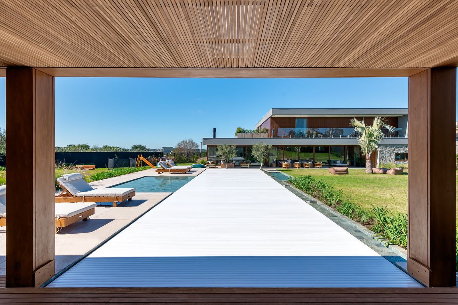 Projeto paisagístico contemporâneo de grande porte integrado a uma residência de arquitetura moderna em dois pavimentos, com piscina retangular alongada equipada com cobertura automática retrátil, emoldurada por um imponente pergolado em madeira ripada que cria um pórtico de contemplação. O jardim combina espécies tropicais e mediterrâneas — oliveiras, palmeiras-leque e capins ornamentais — dispostas em canteiros lineares que acompanham a geometria da piscina, sobre amplo gramado de esmeralda qu, estilo Moderno, Tropical, elementos Piscina retangular de borda infinita com cobertura automática retrátil branca, Pergolado em madeira com laminado ripado no teto, Espreguiçadeiras de madeira com estofados cinza claro, Muro perimetral escuro com cerca de segurança no topo, Casa principal de dois pavimentos com fachada em madeira, vidro e pedra natural, Playground infantil com escorregador amarelo ao fundo, Deck em concreto ao redor da piscina, Canteiros de plantio entre a piscina e o gramado, Pufes e mobiliário lounge na área coberta da residência, Lareira externa com chaminé metálica visível na fachada, plantas Palmeira-leque-da-china, Oliveira, Capim-do-texas, Moréia, Agapanto, Grama-esmeralda, Formio, Aspargo-pluma, ambiente sofisticado, moderno, relaxante, localização piscina, jardim, quintal, área grande, estruturas pergolado, churrasqueira, iluminacao, banco, cores verde, branco, marrom, cinza, bege, amarelo, azul-céu, piso grama, pedra, porcelanato, privacidade cerca_viva, muro_verde, água piscina