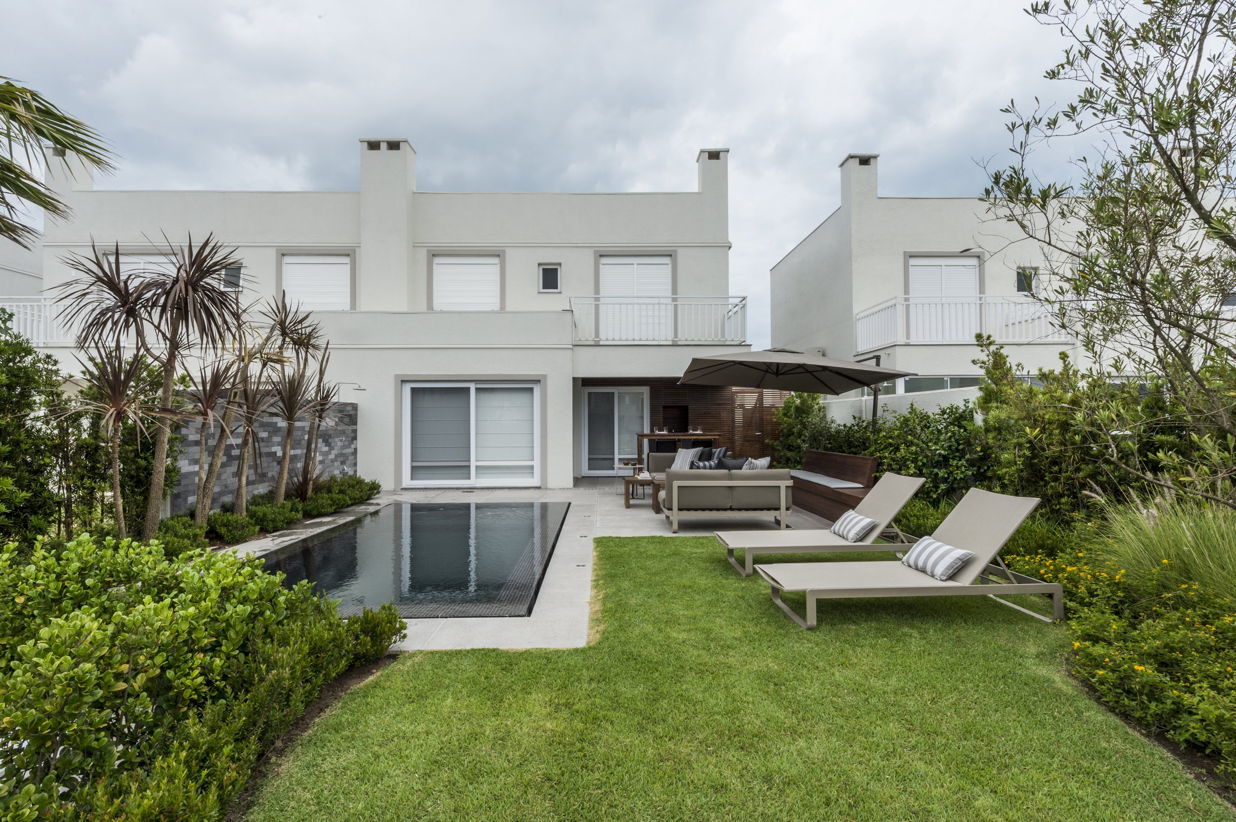 Projeto paisagístico contemporâneo integrado a uma residência de dois pavimentos com linhas retas e acabamento em tons neutros, onde a piscina de borda infinita com revestimento escuro funciona como espelho d'água central, ladeada por uma parede revestida em pedra natural cinza que confere textura e sofisticação. O jardim combina espécies tropicais como dracenas e palmeiras com oliveiras de influência mediterrânea, enquanto a cerca viva de clúsia garante privacidade e o gramado esmeralda cria tr, estilo Moderno, Tropical, elementos Piscina retangular com borda infinita e revestimento escuro, Parede de pedra natural em tons de cinza como cascata decorativa, Espreguiçadeiras em alumínio com almofadas listradas branco e cinza, Sofá e mesa de centro em área lounge coberta por ombrelone escuro, Área gourmet com churrasqueira e painel ripado em madeira, Piso em porcelanato claro ao redor da piscina, Gramado bem aparado em formato orgânico contornando a piscina, Cerca viva de clúsia delimitando laterais do jardim, Balcões com guarda-corpo metálico branco no pavimento superior, plantas Dracena, Clúsia, Oliveira, Buxinho, Grama-esmeralda, Lantana, Palmeira-garrafa, ambiente sofisticado, moderno, relaxante, localização jardim, piscina, quintal, área medio, estruturas churrasqueira, iluminacao, cores verde-escuro, branco, cinza, grafite, bege, marrom, piso grama, porcelanato, pedra, privacidade cerca_viva, água piscina