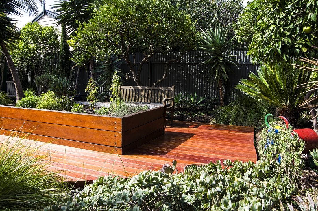 Jardim de quintal residencial com estilo tropical-contemporâneo, organizado em torno de um generoso deck de madeira nobre avermelhada que funciona como área de estar e contemplação, acompanhado por um banco clássico de madeira posicionado junto a uma jardineira elevada com ervas e trepadeiras. O paisagismo combina uma exuberante massa vegetal tropical ao fundo — com palmeiras, dracenas, agaves e árvores frutíferas que garantem privacidade contra a cerca metálica corrugada — com um primeiro plano, estilo Moderno, Tropical, elementos Deck de madeira de lei (aparência de Itaúba ou Cumaru) com acabamento envernizado, Jardineira elevada em madeira de lei com bordas robustas e parafusos aparentes, Banco de jardim clássico em madeira com encosto de ripas, Escultura decorativa em cerâmica/vidro colorido em formato de pássaro/flamingo vermelho, Cerca de chapa metálica corrugada (galvanizada escura) como limite do terreno, Tutor/treliça para planta trepadeira junto à jardineira, Mangueira de jardim verde enrolada ao lado do deck, plantas Capim-do-texas, Planta-jade, Palmeira-leque, Dracena, Agave, Árvore-da-felicidade, Limoeiro, Palmeira-ráfis, Babosa, Strelitzia, Zamioculca, Grama-preta, Suculenta-rosa-de-pedra, Cycas, Aspargo-pluma, ambiente relaxante, acolhedor, sofisticado, localização quintal, jardim, área medio, estruturas banco, cores Verde escuro, Verde claro, Marrom avermelhado (deck), Cinza escuro (cerca metálica), Vermelho (escultura), Azul e multicolorido (escultura cerâmica), Verde acinzentado (suculentas), Amarelo esverdeado (folhagens novas), piso deck, privacidade cerca_viva, muro_verde, água nenhum