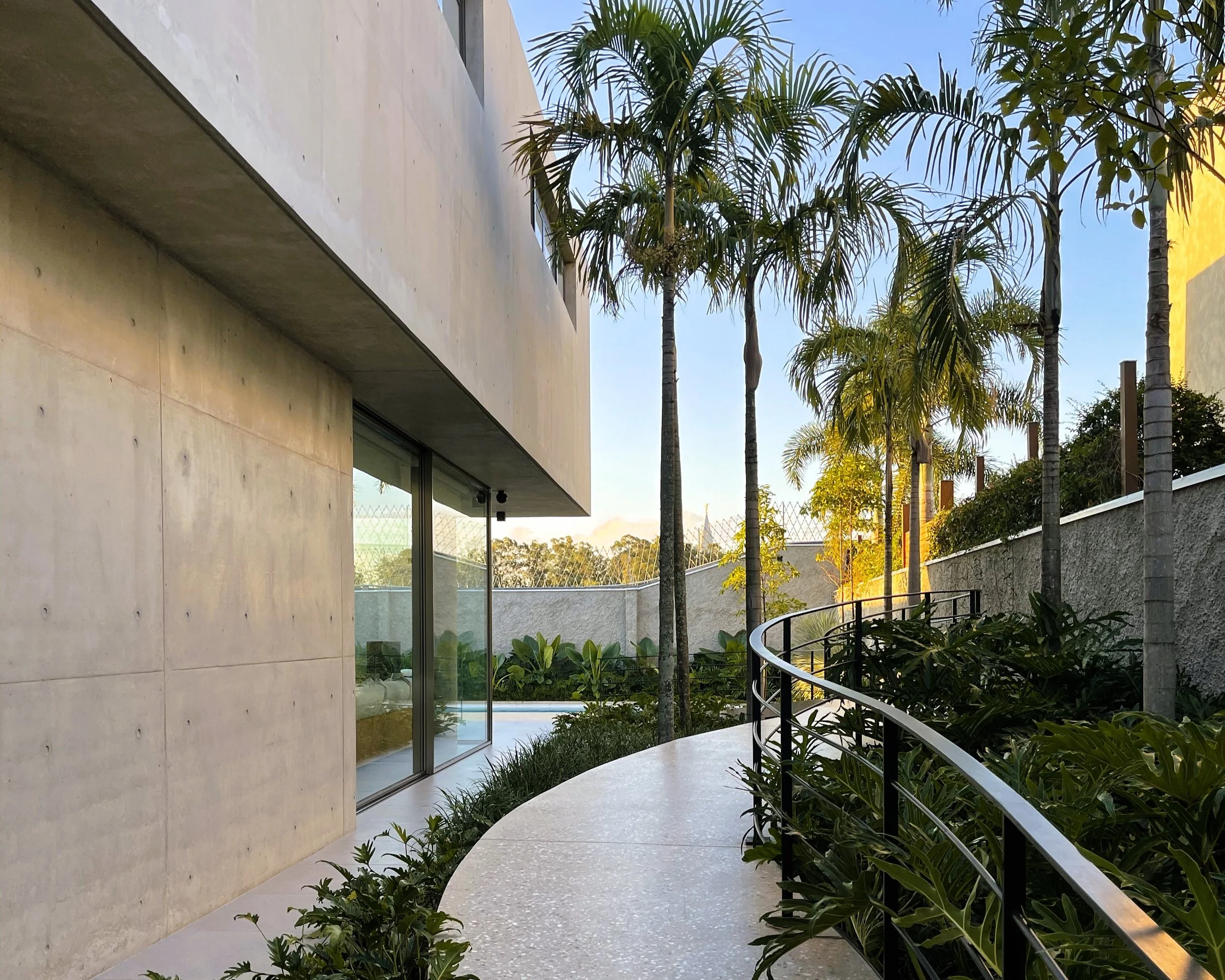 Projeto paisagístico contemporâneo integrado a uma residência de concreto aparente, onde uma passarela curva em concreto polido com agregados expostos serpenteia entre canteiros tropicais exuberantes compostos por palmeiras-reais de grande porte, filodendros, costelas-de-adão e forrações de barba-de-serpente. O guarda-corpo metálico em aço preto com desenho curvilíneo acompanha organicamente o traçado da passarela, criando uma transição elegante entre o volume arquitetônico brutalista e o jardim, estilo Moderno, Tropical, elementos Passarela curva em concreto polido com agregados expostos, Guarda-corpo metálico curvo em aço preto com perfil tubular, Parede em concreto aparente com furos de forma (tie holes), Esquadria piso-teto em alumínio preto com vidro amplo, Muro de divisa em concreto/alvenaria revestido, Canteiro perimetral com vegetação tropical densa, Volume superior em balanço (concreto aparente), Tela de proteção diamantada sobre muro, plantas Palmeira-real, Costela-de-adão, Barba-de-serpente, Filodendro, Pacová, Aglaonema, Lírio-da-paz, ambiente sofisticado, moderno, relaxante, localização corredor, jardim, piscina, fachada, área grande, estruturas iluminacao, cores Cinza concreto, Verde escuro, Verde claro, Preto, Azul céu, Branco, Dourado (luz solar), piso porcelanato, pedra, privacidade muro_verde, cerca_viva, água piscina
