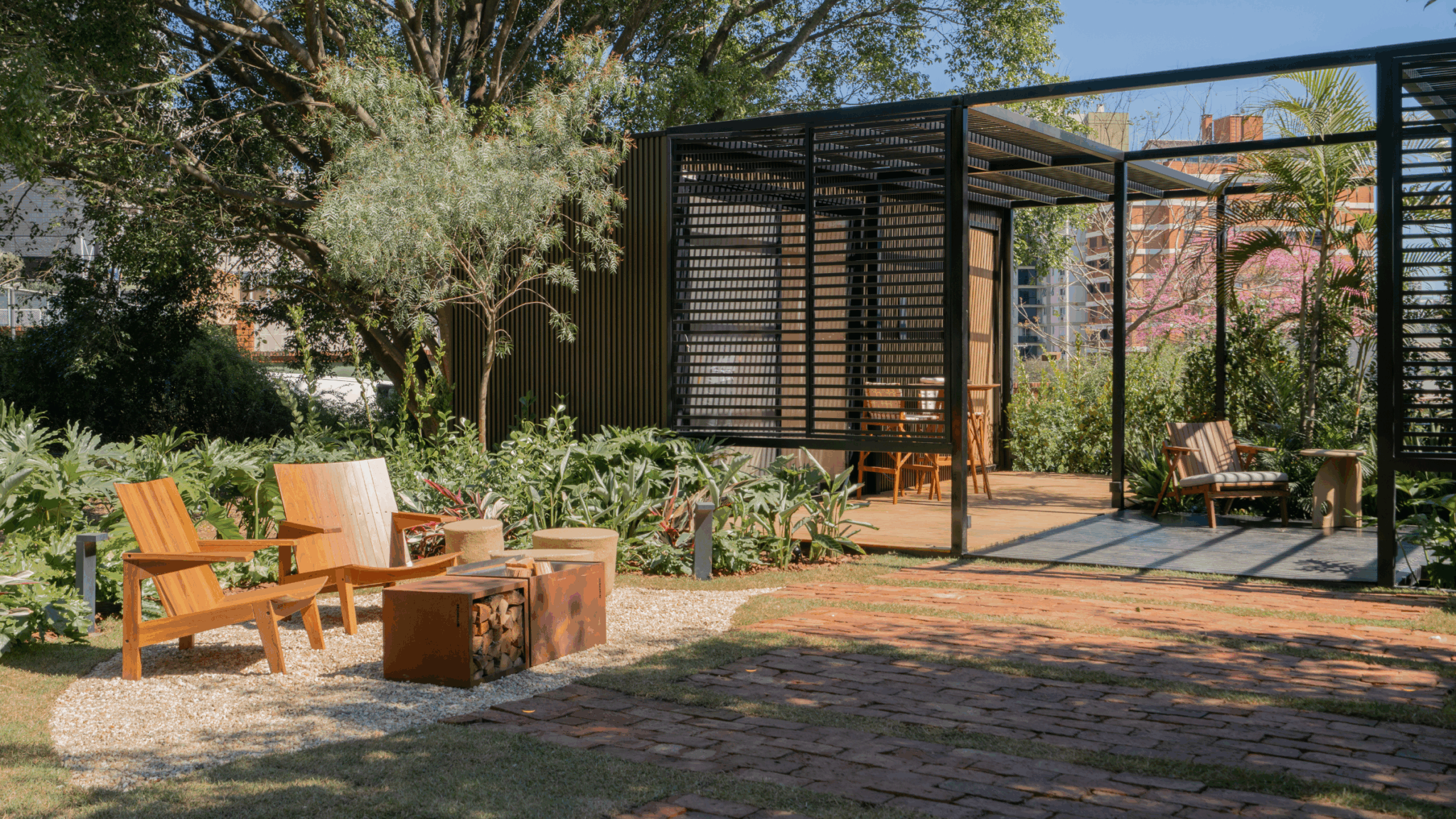 Jardim contemporâneo brasileiro de médio porte que combina uma robusta estrutura de pergolado em aço preto com brises metálicos horizontais, criando uma área de estar sombreada com deck de madeira, integrada a um lounge externo com poltronas Adirondack em teca dispostas ao redor de um elegante fire pit em aço corten com depósito de lenha embutido. O paisagismo tropical exuberante, composto por maciços de filodendros, samambaias, helicônias e antúrios, envolve organicamente as estruturas rígidas,, estilo Moderno, Tropical, elementos Poltronas Adirondack em madeira maciça teca, Fogueira/fire pit em aço corten com compartimento para lenha, Mesinha lateral em concreto ou pedra natural cilíndrica, Pergolado estrutural em metalon de aço preto com brises horizontais, Deck em madeira natural sob o pergolado, Cadeiras de design em madeira maciça sob o pergolado, Piso em tijolo de demolição assentado em padrão espinha de peixe, Área de brita/pedrisco claro ao redor da área de estar, Balizadores de iluminação em aço preto ao longo do caminho, Painel vertical em ripas metálicas pretas como pano de fundo, Almofada listrada sobre cadeira lateral, plantas Samambaia-açu, Filodendro, Helicônia, Costela-de-adão, Oliveira, Ipê-rosa, Palmeira-real, Asplênio, Aglaonema, Antúrio, Grama-esmeralda, Jabuticabeira ou árvore nativa de grande porte, ambiente relaxante, moderno, sofisticado, acolhedor, localização jardim, quintal, área medio, estruturas pergolado, iluminacao, banco, cores Verde intenso, Preto fosco, Madeira natural/teca, Terracota/aço corten, Bege/areia, Rosa (florada do ipê), Marrom tijolo, piso grama, deck, brita, pisantes, privacidade cerca_viva, muro_verde, água nenhum