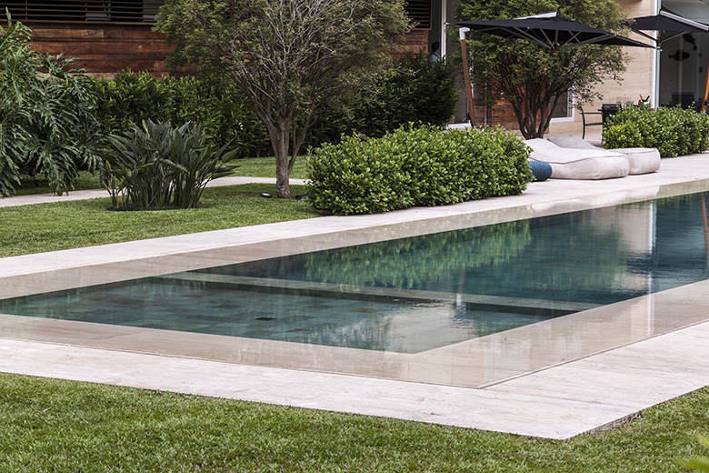 Projeto paisagístico contemporâneo de alto padrão com piscina de borda infinita revestida em tons escuros e bordas em mármore travertino romano, integrada a um amplo gramado de grama-esmeralda impecavelmente mantido. A volumetria vegetal é composta por maciços de buxinho podados em formas orgânicas arredondadas junto à borda da piscina, jabuticabeiras de tronco escultórico como ponto focal e helicônias tropicais adensando os canteiros junto ao muro de madeira. A área de descanso com almofadões d, estilo Moderno, Tropical, elementos piscina com borda infinita e degrau interno em tom escuro, bordas de piscina em mármore travertino, almofadões decorativos em tons neutros e azul, ombrelone lateral preto de braço articulado, fachada com revestimento em madeira e venezianas, canteiro elevado junto ao muro com vegetação tropical, topiaria de buxinho em formato arredondado junto à borda da piscina, plantas Buxinho, Helicônia, Jabuticabeira, Grama-esmeralda, Palmeira-ráfis, ambiente sofisticado, moderno, relaxante, localização piscina, jardim, área grande, estruturas iluminacao, cores verde-escuro, verde-claro, bege, azul-petróleo, branco, marrom-madeira, preto, cinza, piso grama, pedra, privacidade cerca_viva, muro_verde, água piscina