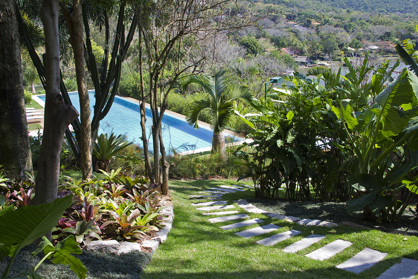 Projeto paisagístico tropical de grande porte implantado em terreno com aclive generoso, onde um caminho de pisantes retangulares de pedra natural serpenteia sobre grama-esmeralda impecável, conduzindo o olhar até uma ampla piscina de borda infinita com revestimento em pastilha azul que se funde visualmente à vegetação exuberante do vale ao fundo. Canteiros elevados delimitados por muretas de pedra natural abrigam maciços de bromélias em tons de vinho e verde, sombreados por dracenas e árvores d, estilo Tropical, Moderno, elementos Pisantes retangulares de pedra natural sobre grama, Piscina de borda infinita com revestimento em pastilha azul, Mureta de pedra natural delimitando canteiro elevado, Espreguiçadeiras em madeira ao redor da piscina, Caminho sinuoso de pisantes conduzindo à área da piscina, Canteiros orgânicos com forrações tropicais, Desnível natural do terreno aproveitado no projeto paisagístico, plantas Bromélia-imperial, Bromélia, Palmeira-leque, Helicônia, Bananeira-ornamental, Dracena, Filodendro, Palmeira-areca, Grama-esmeralda, Grama-preta, Árvore-polvo, Bambu-mossô, Costela-de-adão, Taioba-ornamental, ambiente relaxante, sofisticado, localização jardim, piscina, área grande, estruturas nenhum, cores verde-escuro, verde-claro, vinho, rosa-envelhecido, azul-piscina, cinza-pedra, marrom-terra, bege, piso grama, pisantes, pedra, privacidade cerca_viva, água piscina