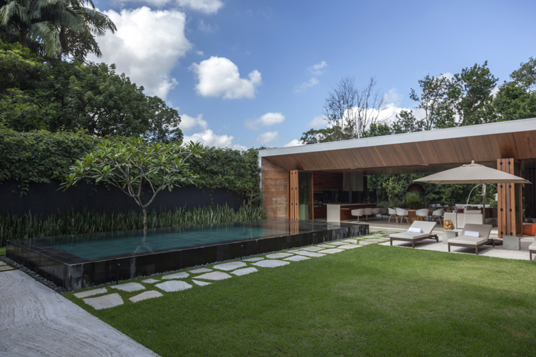 Projeto paisagístico contemporâneo de alto padrão que integra uma piscina de borda infinita com acabamento em pedra escura a um amplo gramado de grama-esmeralda, pontuado por pisantes irregulares de pedra natural que conduzem o percurso. Um exemplar escultural de jasmim-manga (Plumeria rubra) posicionado estrategicamente junto à piscina cria um ponto focal tropical, enquanto o muro escuro ao fundo recebe vegetação densa de helicônias e papiros que garantem privacidade e verticalidade. A área de , estilo Moderno, Tropical, elementos Piscina com borda infinita em acabamento escuro, Espreguiçadeiras em fibra sintética com almofadas brancas, Ombrelone de tecido bege, Pisantes de pedra natural sobre grama, Muro de contenção escuro ao fundo com vegetação alta, Cobertura plana com forro em madeira ripada, Pilares em madeira maciça, Seixos rolados na borda da piscina, Área gourmet integrada ao espaço da piscina, Canteiro elevado junto ao muro, Gramado amplo e bem aparado, plantas Jasmim-manga, Helicônia, Palmeira, Grama-esmeralda, Moréia, Papiro, Mangueira, Pândano, Singônio, ambiente relaxante, moderno, sofisticado, localização jardim, piscina, quintal, área grande, estruturas churrasqueira, pergolado, iluminacao, cores verde-escuro, verde-claro, cinza-escuro, bege, branco, marrom-madeira, azul-esverdeado, piso grama, pisantes, pedra, privacidade muro_verde, cerca_viva, água piscina