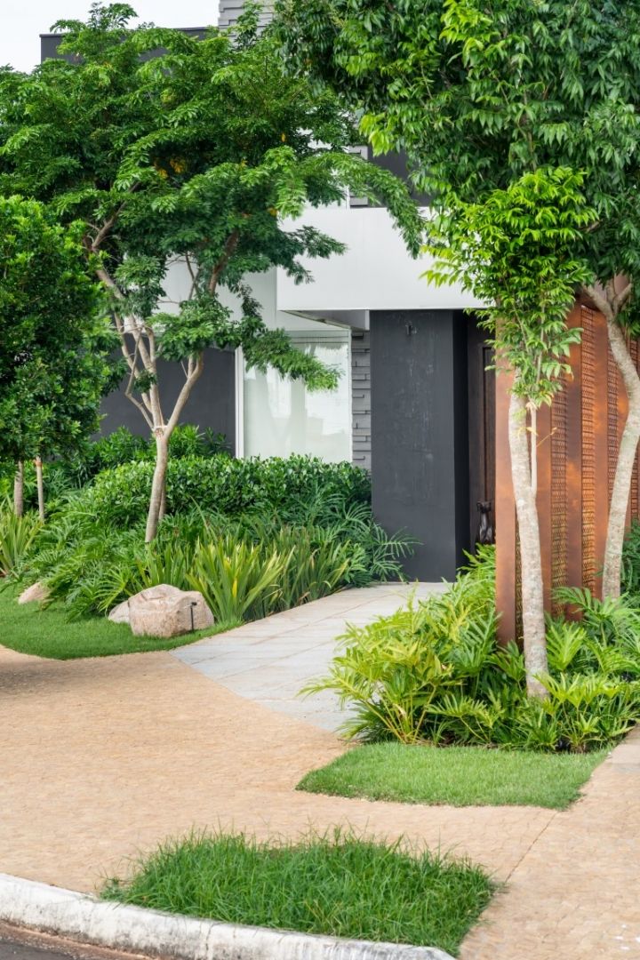 Projeto de paisagismo contemporâneo em fachada residencial que integra um volumoso maciço tropical composto por clusias, filodendros, palmeiras-ráfis e moréias, criando camadas verdes de diferentes alturas e texturas que suavizam a arquitetura em concreto aparente cinza e branco. O caminho de acesso em pisantes de pedra natural em tom areia, intercalados por faixas de grama-esmeralda, conduz o olhar até o imponente portão em aço corten perfurado, que dialoga com os troncos das árvores de médio p, estilo Moderno, Tropical, elementos Painel em aço corten perfurado como portão de entrada, Piso em pisantes de pedra natural tons areia/ocre, Faixas de grama entre pisantes, Pedra natural decorativa sobre a grama, Iluminação de jardim embutida no solo (spot tipo balizador), Fachada em concreto aparente cinza escuro e branco, Janela em vidro fixo ampla, plantas Pata-de-vaca, Clusia, Moréia, Palmeira-ráfis, Grama-esmeralda, Liriope, Filodendro-xanadu, Helicônia, Jabuticabeira, Renda-portuguesa, Aglaonema, Zamioculca, Pleomele, ambiente moderno, sofisticado, acolhedor, localização fachada, entrada, jardim, área medio, estruturas iluminacao, cores Verde intenso, Verde claro, Areia/bege (piso), Ferrugem/corten, Cinza escuro (fachada), Branco (fachada), piso pisantes, grama, privacidade cerca_viva, água nenhum