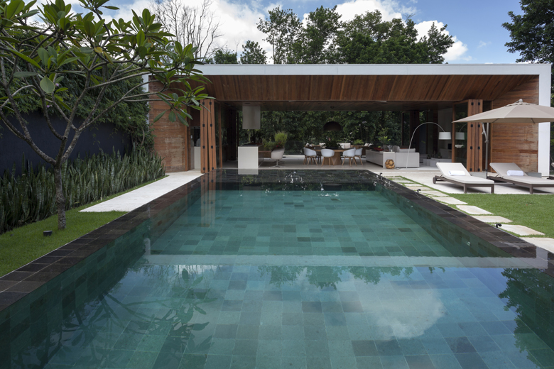 Projeto paisagístico contemporâneo de ampla área de lazer integrada a um pavilhão de estar e jantar aberto, com piscina retangular revestida em pedra Hijau que cria reflexos espetaculares da vegetação e do céu, emoldurada por faixas de grama-esmeralda impecável. A composição botânica combina uma Plumeria adulta como ponto focal escultural à esquerda, com massas de espadas-de-são-jorge e agaves em canteiro junto ao muro escuro, criando forte contraste cromático e textural. A arquitetura em taipa , estilo Moderno, Tropical, elementos Piscina retangular com revestimento em pedra Hijau (tons de verde), Borda de piscina em pedra natural escura, Pavilhão com cobertura plana em laje branca e forro de madeira, Pilares e painéis em taipa de pilão/tijolo aparente, Espreguiçadeiras de madeira com estofado branco, Ombrelone bege de braço lateral, Mobiliário de estar externo com sofás brancos e mesa de jantar com cadeiras Eames brancas, Muro de divisa pintado em preto/chumbo, Pisantes de pedra clara entre a grama, Canteiro elevado lateral junto ao muro, plantas Plumeria, Espada-de-são-jorge, Agave, Palmeira-leque, Helicônia, Costela-de-adão, Grama-esmeralda, Philodendro, Clusia, ambiente relaxante, moderno, sofisticado, localização piscina, jardim, quintal, área grande, estruturas gazebo, iluminacao, cores verde-esmeralda, branco, madeira natural, terracota, cinza-chumbo, bege, preto, piso grama, pedra, pisantes, privacidade muro_verde, cerca_viva, água piscina