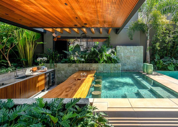 Projeto paisagístico tropical contemporâneo que integra magistralmente uma piscina com revestimento em pedra natural esverdeada a uma sofisticada área gourmet, conectadas por uma bancada-bar em madeira maciça com borda orgânica viva suspensa sobre a água, acompanhada de bancos submersos em pedra que permitem a experiência de swim-up bar. A vegetação exuberante composta por palmeiras, helicônias, filodendros e costelas-de-adão cria uma atmosfera de selva tropical envolvente, emoldurada pelo forro, estilo Moderno, Tropical, elementos Piscina revestida em pedra natural tom esverdeado, Bancada-bar em madeira maciça com borda orgânica suspensa sobre a piscina, Bancos submersos em pedra dentro da piscina tipo swim-up bar, Parede de pedra natural com textura rústica ao fundo, Forro em madeira ripada tom freijó/cumaru na cobertura, Cozinha gourmet externa com gabinete em madeira ripada, Ventilador de teto estilo colonial em bronze, Luminárias pendentes tipo gaiola em fibra natural, Cadeira suspensa tipo balanço em rattan/vime, Vaso cerâmico grande em tom esverdeado, Taças de vinho sobre a bancada do bar, Degraus em pedra natural para acesso à piscina, Estrutura metálica aparente em aço escuro com vigas de madeira, plantas Helicônia, Palmeira-areca, Costela-de-adão, Filodendro, Lírio-da-paz, Bananeira-ornamental, Pacová, Singônio, Palmeira-rápis, Antúrio, Orquídea, Jiboia, Samambaia, Taioba-ornamental, Palmeira-real, Calateia, ambiente relaxante, moderno, sofisticado, localização piscina, quintal, jardim, área medio, estruturas pergolado, churrasqueira, iluminacao, cores Verde-escuro, Verde-esmeralda (água da piscina), Madeira mel/cumaru, Cinza-chumbo, Bege-areia, Branco, Bronze, piso pedra, deck, privacidade muro_verde, cerca_viva, água piscina