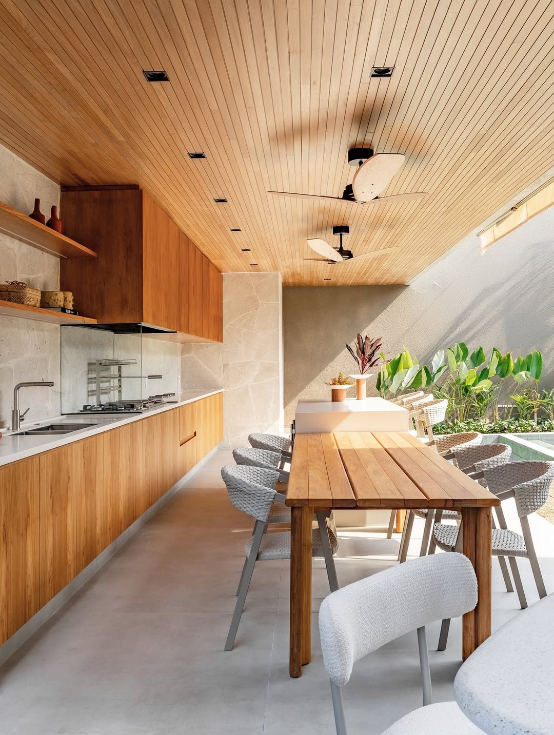 Espaço gourmet semi-coberto com linguagem arquitetônica contemporânea tropical, integrando cozinha linear com bancada em concreto polido e marcenaria em teca sob um generoso forro em réguas de madeira natural equipado com ventiladores de teto. A mesa de jantar em madeira maciça para aproximadamente 8 pessoas posiciona-se estrategicamente voltada para o jardim lateral com vegetação tropical exuberante de bananeiras-ornamentais e helicônias que emolduram a borda da piscina. O projeto paisagístico , estilo Moderno, Tropical, elementos mesa de jantar em madeira teca maciça com acabamento natural, cadeiras de jantar em fibra sintética trançada cinza com estrutura metálica, cadeira estofada em bouclé branco com pés metálicos, cozinha gourmet com bancada em concreto polido, armários e gabinetes em madeira teca com puxadores embutidos, forro em réguas de madeira teca com acabamento natural, ventiladores de teto com pás em madeira escura, spots de embutir quadrados no forro, prateleiras em madeira na parede da cozinha, parede revestida em pedra natural tipo palimanan, splashback em vidro transparente na área do cooktop, vaso decorativo em cerâmica com planta vermelha, objetos decorativos em cerâmica terracota nas prateleiras, piso em cimento queimado claro, abertura lateral com vista para a piscina e jardim tropical, plantas Helicônia, Bananeira-ornamental, Filodendro, Dracena-vermelha, ambiente relaxante, moderno, sofisticado, acolhedor, localização varanda, piscina, quintal, área medio, estruturas churrasqueira, iluminacao, cores madeira natural (tons de teca dourada), cinza claro (concreto e piso), bege arenoso (pedra natural), verde vibrante (folhagens tropicais), branco (estofados e bancada), terracota (objetos decorativos), verde-água (piscina ao fundo), piso porcelanato, privacidade muro_verde, água piscina