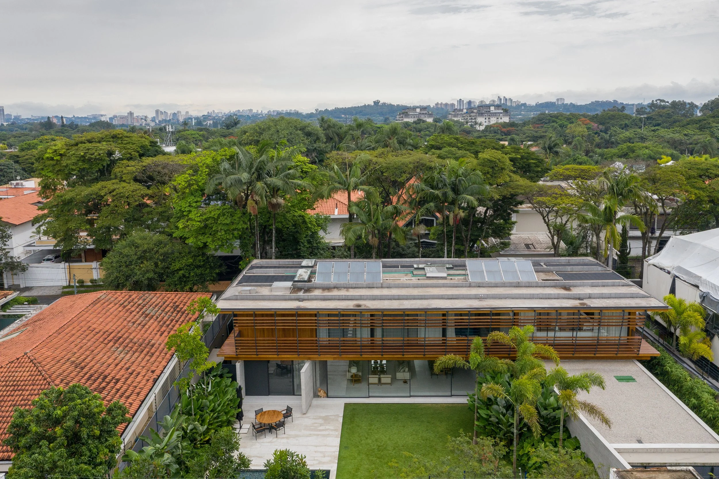 Residência contemporânea de alto padrão em bairro arborizado de São Paulo, com volumetria marcada por um pavimento superior revestido em brises de madeira cumaru em balanço sobre base de concreto aparente e extensos panos de vidro, integrando os ambientes internos ao jardim tropical circundante. O paisagismo exuberante combina palmeiras-reais, arecas, bananeiras-ornamentais e estrelítzias em canteiros perimetrais que criam privacidade natural, enquanto o gramado esmeralda central e o piso de por, estilo Moderno, Tropical, elementos Painéis solares fotovoltaicos no telhado plano, Brises de madeira ripada na fachada superior, Laje em balanço de concreto aparente, Panos de vidro piso-teto no pavimento térreo, Mesa de jantar externa em madeira com cadeiras, Piso de porcelanato claro no pátio externo, Gramado central bem aparado, Canteiros tropicais perimetrais, Cobertura plana com platibanda em concreto, Pilares de concreto aparente sustentando o volume superior, plantas Palmeira-real, Palmeira-areca, Helicônia, Bananeira-ornamental, Palmeira-rápis, Estrelítzia-branca, Pau-brasil, Jabuticabeira, Mangueira, Ficus, Filodendro, Grama-esmeralda, Clúsia, Palmeira-imperial, Sibipiruna, ambiente moderno, sofisticado, relaxante, localização jardim, fachada, quintal, varanda, entrada, área grande, estruturas iluminacao, banco, cores Verde intenso das copas tropicais, Marrom-dourado dos brises de madeira, Cinza do concreto aparente, Branco/bege do porcelanato, Verde-claro do gramado, Terracota dos telhados vizinhos, Azul-escuro dos painéis solares, piso grama, porcelanato, privacidade cerca_viva, água nenhum