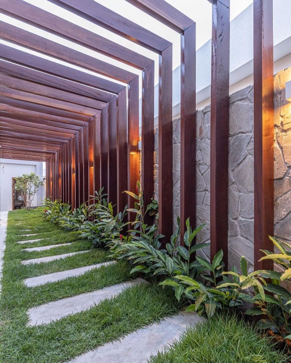 Corredor lateral residencial contemporâneo com pergolado estruturado em perfis de aço corten dispostos ritmicamente em lâminas verticais e travessas horizontais, criando um jogo de luz e sombra ao longo do percurso. O muro de pedra natural irregular em tons de cinza é valorizado por arandelas de luz quente embutidas entre os perfis metálicos, enquanto um canteiro linear de helicônias, pacovás e clúsias confere exuberância tropical ao conjunto. Pisantes retangulares sobre grama-esmeralda compõem , estilo Moderno, Tropical, elementos Pergolado em aço corten com lâminas verticais e horizontais repetidas em ritmo linear, Muro de pedra natural irregular (pedra madeira ou pedra moledo) em tons de cinza, Pisantes retangulares de pedra natural ou concreto embutidos na grama, Iluminação de parede embutida tipo arandela com facho bidirecional em luz quente âmbar, Canteiro linear junto ao muro com forração tropical, Grama entre os pisantes como piso permeável, plantas Helicônia, Pacová, Clúsia, Grama-esmeralda, Pleomele, Plumeria, ambiente sofisticado, moderno, acolhedor, localização corredor, jardim, muro, área medio, estruturas pergolado, iluminacao, cores Marrom-ferrugem do aço corten, Cinza da pedra natural, Verde intenso da vegetação, Verde-claro da grama, Âmbar da iluminação, Branco da parede ao fundo, piso grama, pisantes, pedra, privacidade muro_verde, cerca_viva, água nenhum