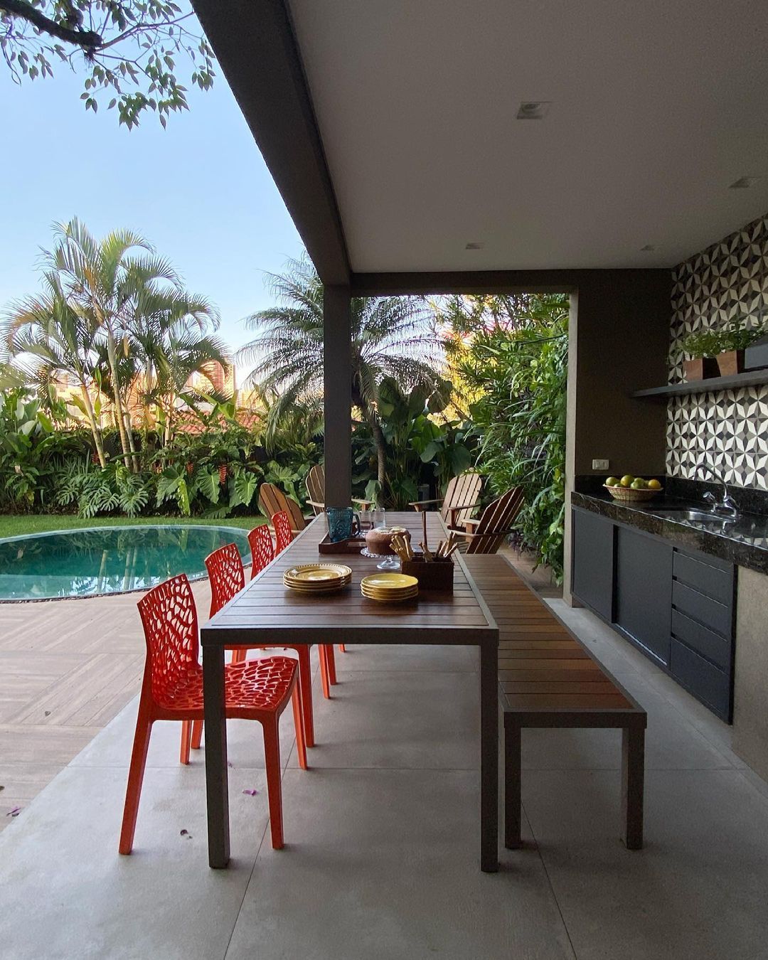 Área de lazer com linguagem contemporânea e paisagismo tropical exuberante que integra piscina de borda orgânica com espaço gourmet coberto, emoldurada por maciços densos de palmeiras, heliconias e filodendros que garantem privacidade e sombreamento natural. A cozinha externa conta com revestimento em ladrilho hidráulico geométrico P&B, bancada em granito escuro e armários grafite, contrastando com as cadeiras vermelhas de design vazado e a madeira quente da mesa e do deck. O conjunto transmite , estilo Moderno, Tropical, elementos Mesa retangular de alumínio com tampo ripado em madeira, Cadeiras vermelhas de polipropileno com design vazado estilo Gruvyer, Banco em madeira com estrutura metálica, Cadeiras Adirondack em madeira natural, Cozinha gourmet externa com bancada em granito escuro, Armários inferiores em tom grafite, Parede revestida com ladrilho hidráulico geométrico preto e branco, Pratos decorativos amarelos sobre a mesa, Cesto com frutas verdes na bancada, Piscina de formato orgânico com borda arredondada, Deck em madeira cumaru ao redor da piscina, Cobertura em laje plana com forro branco e iluminação embutida, Pilar de concreto aparente, Piso em porcelanato grande formato tom bege, plantas Palmeira-imperial, Palmeira-areca, Helicônia, Bananeira-ornamental, Costela-de-adão, Filodendro, Pacová, Grama-esmeralda, Singônio, Samambaia, ambiente relaxante, moderno, sofisticado, acolhedor, localização piscina, varanda, quintal, área medio, estruturas churrasqueira, iluminacao, banco, cores vermelho, verde, bege, grafite, branco, preto, madeira natural, azul turquesa, amarelo, piso porcelanato, deck, grama, privacidade cerca_viva, água piscina