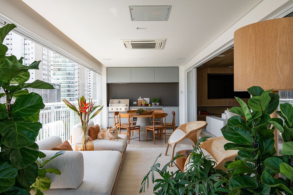 Varanda gourmet de apartamento em andar alto de edifício urbano, integrando área de estar com sofá e poltronas em palhinha a uma zona de refeições com mesa redonda e bancada com churrasqueira a gás, tudo protegido por cortina de vidro retrátil. O paisagismo tropical é protagonizado por exemplares exuberantes de Ficus lyrata posicionados estrategicamente nas extremidades, criando enquadramento verde que suaviza a vista da cidade, complementados por helicônias vermelhas em vaso alto que adicionam , estilo Moderno, Tropical, elementos sofá estofado em linho claro com almofadas em tons terrosos, par de poltronas em madeira com tela de palhinha natural, mesa de jantar redonda com base em concreto e tampo em madeira, cadeiras de jantar em madeira maciça com design autoral, bancada gourmet em granito cinza com armários superiores cinza claro, churrasqueira a gás embutida na bancada, painel de TV em madeira freijó com nicho embutido, luminária pendente cilíndrica em lâmina de madeira, tapete em fibra natural sisal bege, vasos grandes em fibra de vidro ou cimento para as figueiras-lira, fechamento em vidro retrátil tipo cortina de vidro, ar-condicionado tipo cassete embutido no forro, guarda-corpo metálico branco com vidro, plantas Figueira-lira, Helicônia, Pacová, ambiente relaxante, moderno, acolhedor, sofisticado, localização varanda, área medio, estruturas churrasqueira, iluminacao, cores branco, cinza, bege, verde, madeira natural, terracota, piso porcelanato, privacidade nenhum, água nenhum