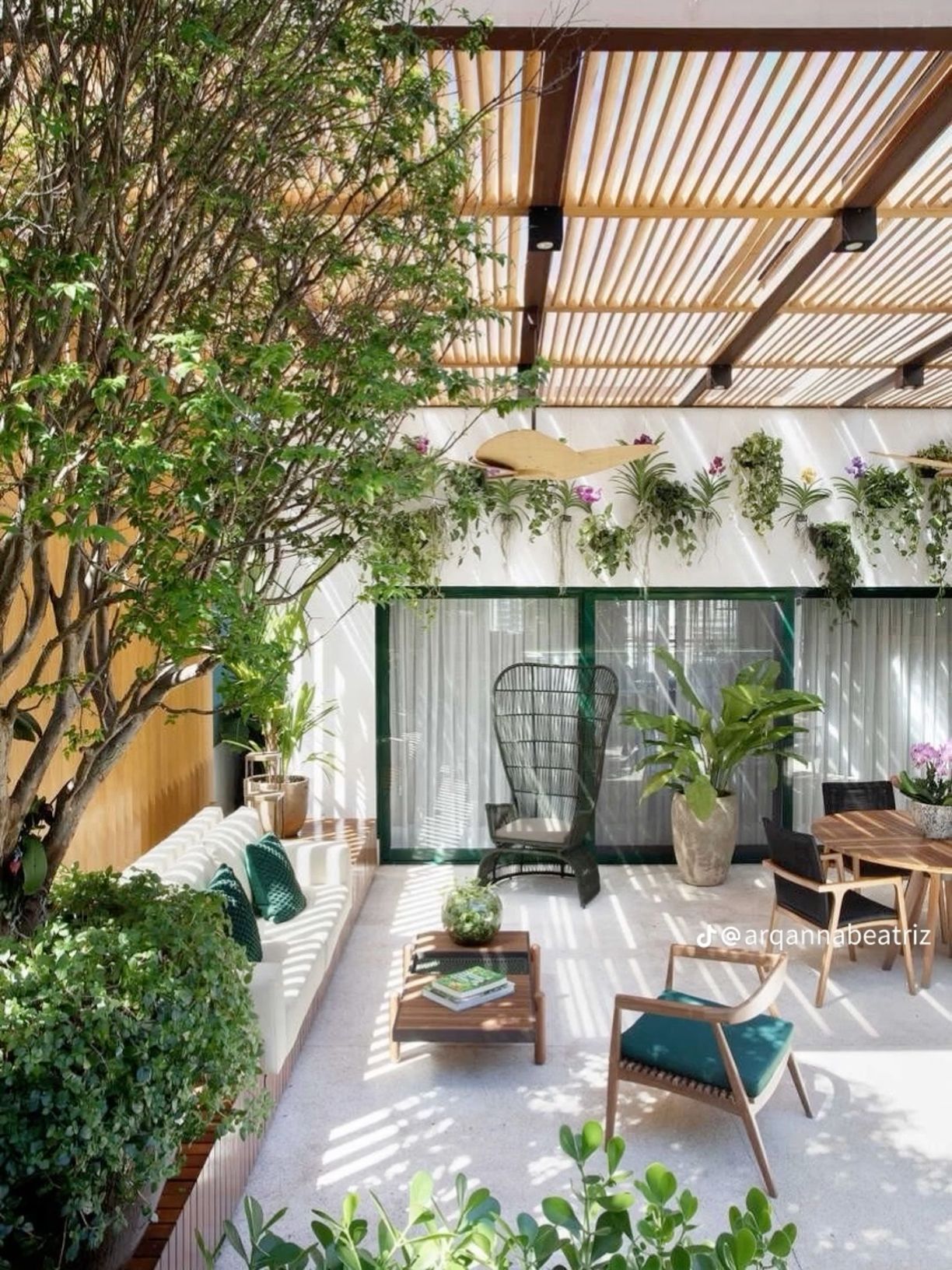 Projeto de área externa semi-coberta com linguagem contemporânea e forte apelo tropical, onde um elegante pergolado de ripas de madeira clara sobre estrutura metálica preta filtra a luz solar criando jogos de sombras lineares sobre o piso de porcelanato claro. O jardim vertical com orquídeas, bromélias e samambaias na parede branca ao fundo compõe com a árvore de médio porte (possivelmente jabuticabeira) plantada em canteiro elevado, criando um microclima agradável e acolhedor. O mobiliário cura, estilo Moderno, Tropical, elementos Pergolado com ripas de madeira clara (freijó ou pinus tratado) sobre estrutura metálica preta, Banco de alvenaria embutido com encosto ripado branco e almofadas verdes esmeralda, Mesa de centro em madeira teca com base metálica preta, Cadeira de balanço tipo Peacock em fibra sintética preta, Poltrona baixa em madeira com assento em tecido verde esmeralda, Mesa lateral de madeira teca com cadeiras de encosto em corda preta, Vasos de parede em meia-lua brancos com jardim vertical, Ventilador de teto decorativo com pá em madeira formato folha, Spots de iluminação embutidos na estrutura do pergolado, Esquadrias de vidro com perfis em alumínio verde escuro e cortinas brancas, Floreira elevada em madeira com forrações, Painel de madeira ripada na lateral esquerda, Vaso grande em cerâmica rústica bege com folhagem tropical, plantas Clúsia, Jabuticabeira, Bromélia, Orquídea, Chifre-de-veado, Pacová, Renda-portuguesa, Singônio, Terrário com musgos e suculentas variadas, Banana-de-jardim, Lírio-da-paz, Jiboia, Asplenium, ambiente relaxante, moderno, sofisticado, acolhedor, localização varanda, corredor, quintal, área medio, estruturas pergolado, banco, iluminacao, cores branco, verde esmeralda, madeira clara (freijó), preto, bege, verde escuro, piso porcelanato, privacidade muro_verde, água nenhum