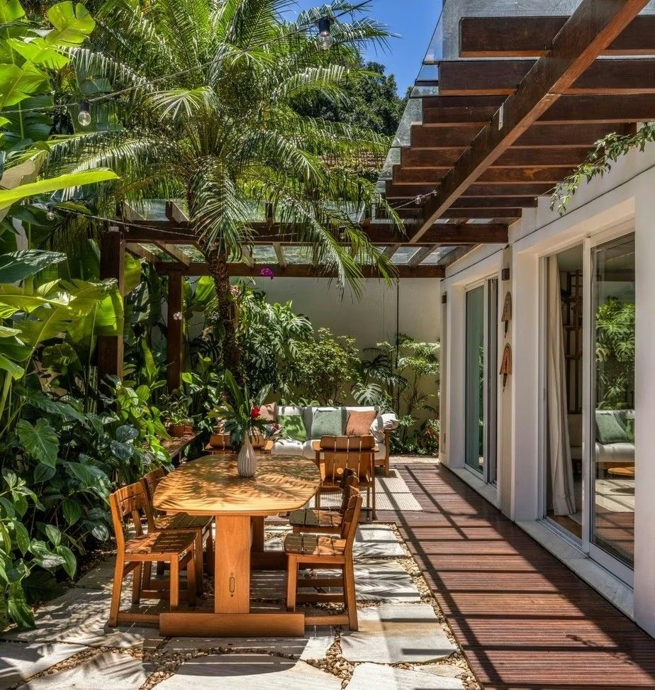 Projeto paisagístico de quintal residencial com forte identidade tropical brasileira, onde um generoso pergolado de madeira escura conecta a área social externa — composta por mesa de jantar em madeira maciça e lounge com sofás estofados — à fachada da casa, integrando interior e exterior por meio de amplas portas de correr em vidro. A vegetação exuberante de porte médio a grande, com palmeiras-reais, bananeiras-ornamentais, costelas-de-adão e helicônias, forma uma cortina verde densa nas latera, estilo Moderno, Tropical, elementos Mesa de jantar externa em madeira maciça com design oval, Cadeiras de madeira com encosto vazado estilo contemporâneo, Sofá externo com estofado branco e almofadas verdes e estampadas, Poltronas de madeira ripada com design moderno, Vaso decorativo branco sobre a mesa, Vasinhos de plantas sobre a mesa, Cordão de luzes (varal de lâmpadas) suspenso no pergolado, Portas de correr em vidro com esquadria branca de alumínio, Cortina branca no vão da porta, Deck de madeira elevado junto à fachada, Pisantes de pedra natural com juntas de seixos rolados, Pergolado de madeira escura com vigas robustas fixado à alvenaria, Mureta ou banco de alvenaria ao fundo com função de apoio, Adorno de parede tipo artesanal junto à porta, plantas Palmeira-real, Helicônia, Costela-de-adão, Bananeira-ornamental, Filodendro, Palmeira-areca, Singônio, Bougainvillea, Lírio-da-paz, Jiboia, Antúrio, ambiente relaxante, acolhedor, sofisticado, localização quintal, varanda, área medio, estruturas pergolado, iluminacao, cores verde-escuro, verde-claro, marrom-madeira, branco, bege, cinza-pedra, rosa-bougainvillea, piso deck, pisantes, brita, pedra, privacidade muro_verde, cerca_viva, água nenhum